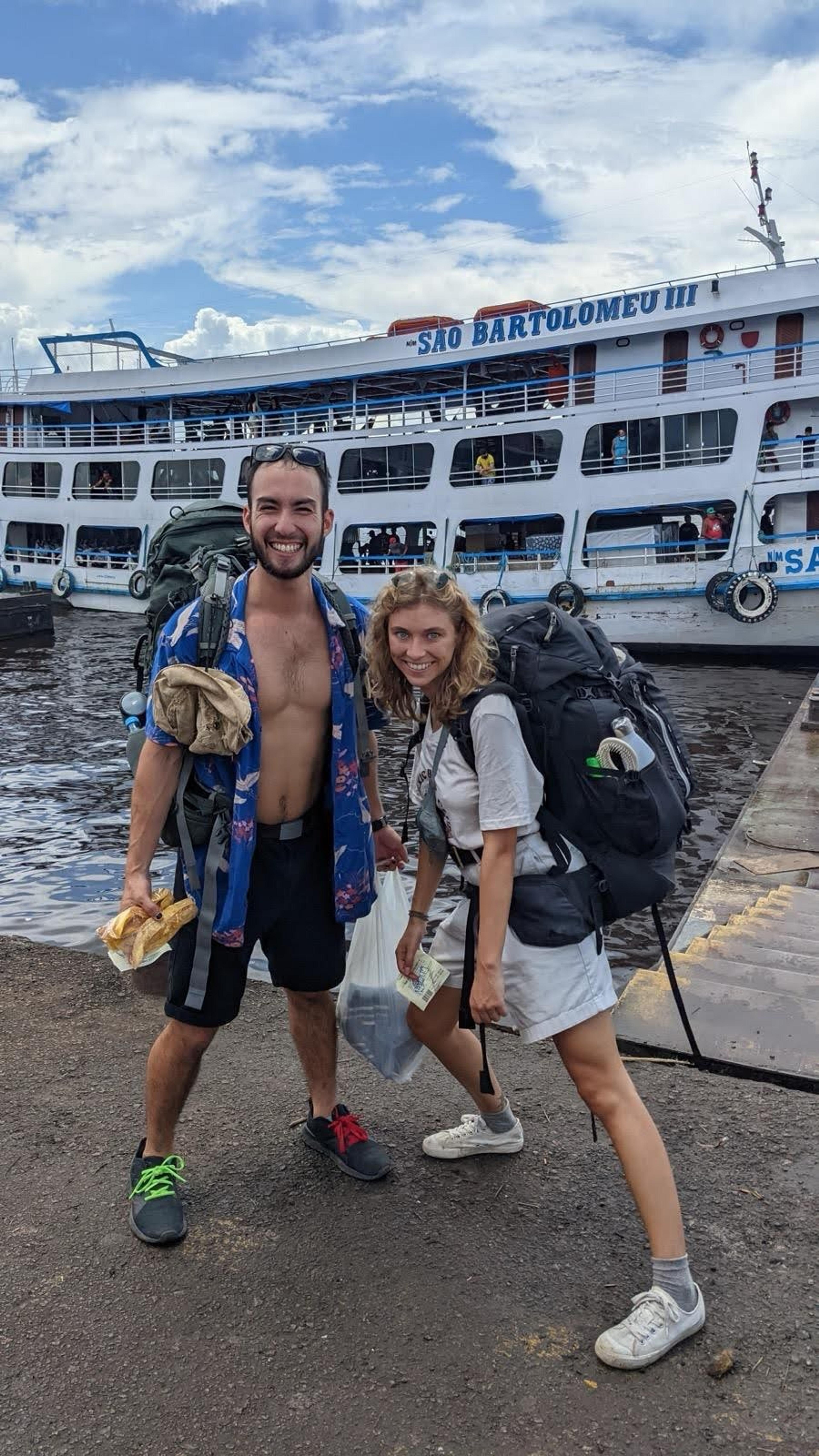 Two travelers with backpacks stand smiling in front of the São Bartolomeu III boat, likely in a harbor setting.