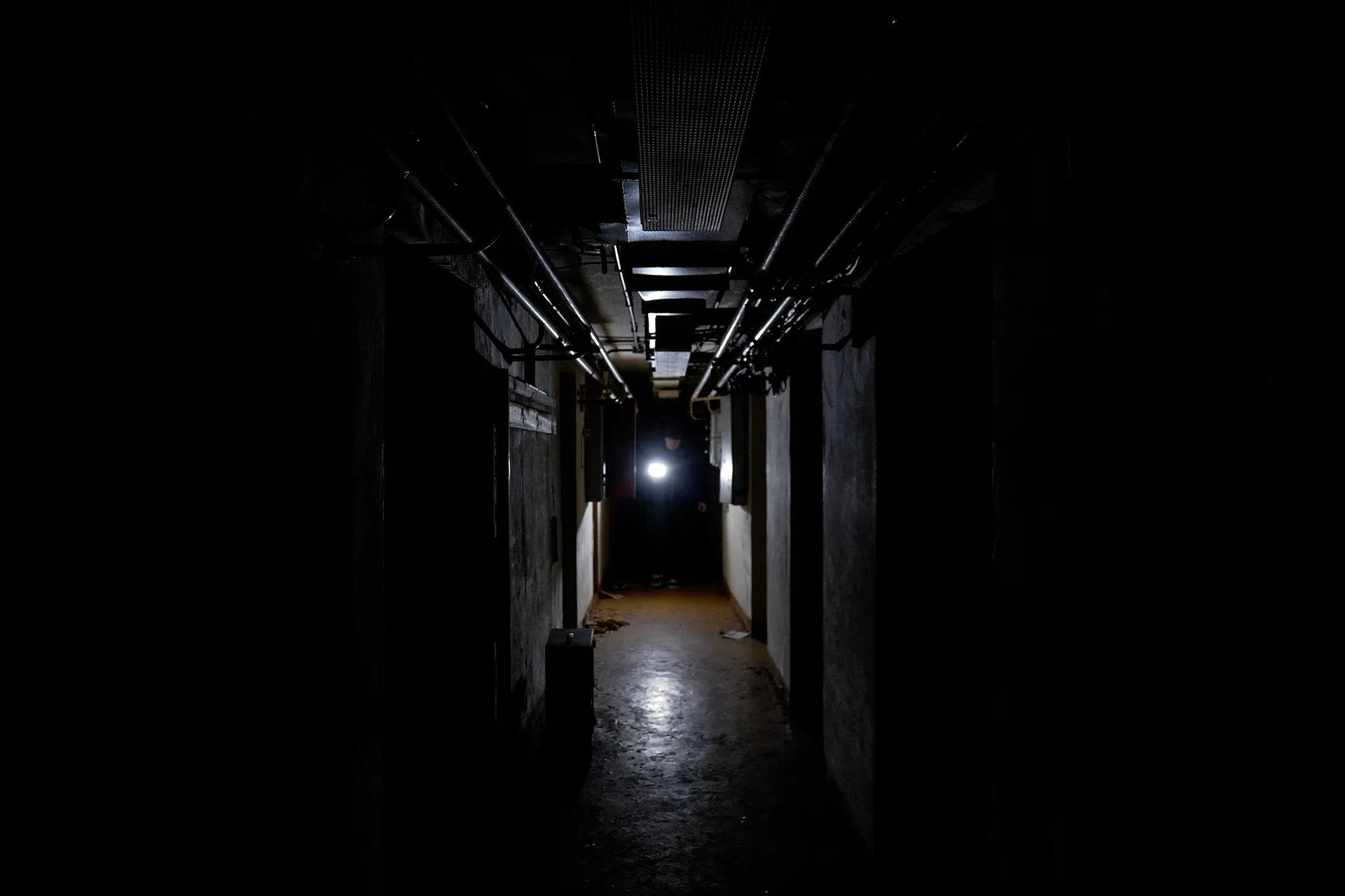 A dimly lit, narrow corridor with exposed pipes overhead and a person holding a flashlight in the distance.