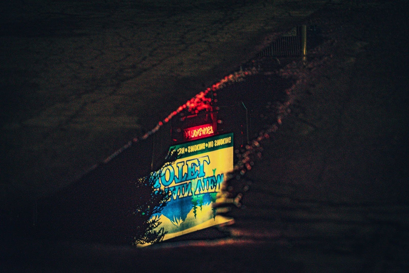 A reflected neon sign with the word "Motel" glows vibrantly in a puddle on a dark street, possibly in an urban area.