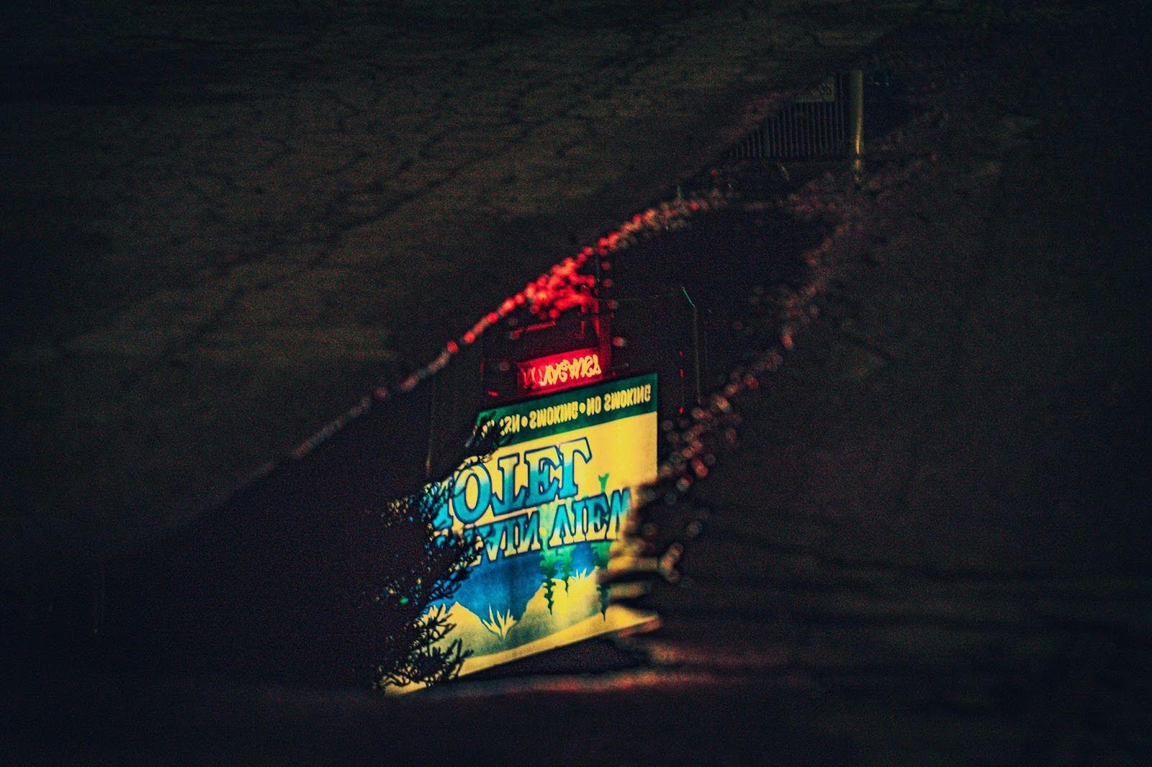 A reflected neon sign with the word "Motel" glows vibrantly in a puddle on a dark street, possibly in an urban area.