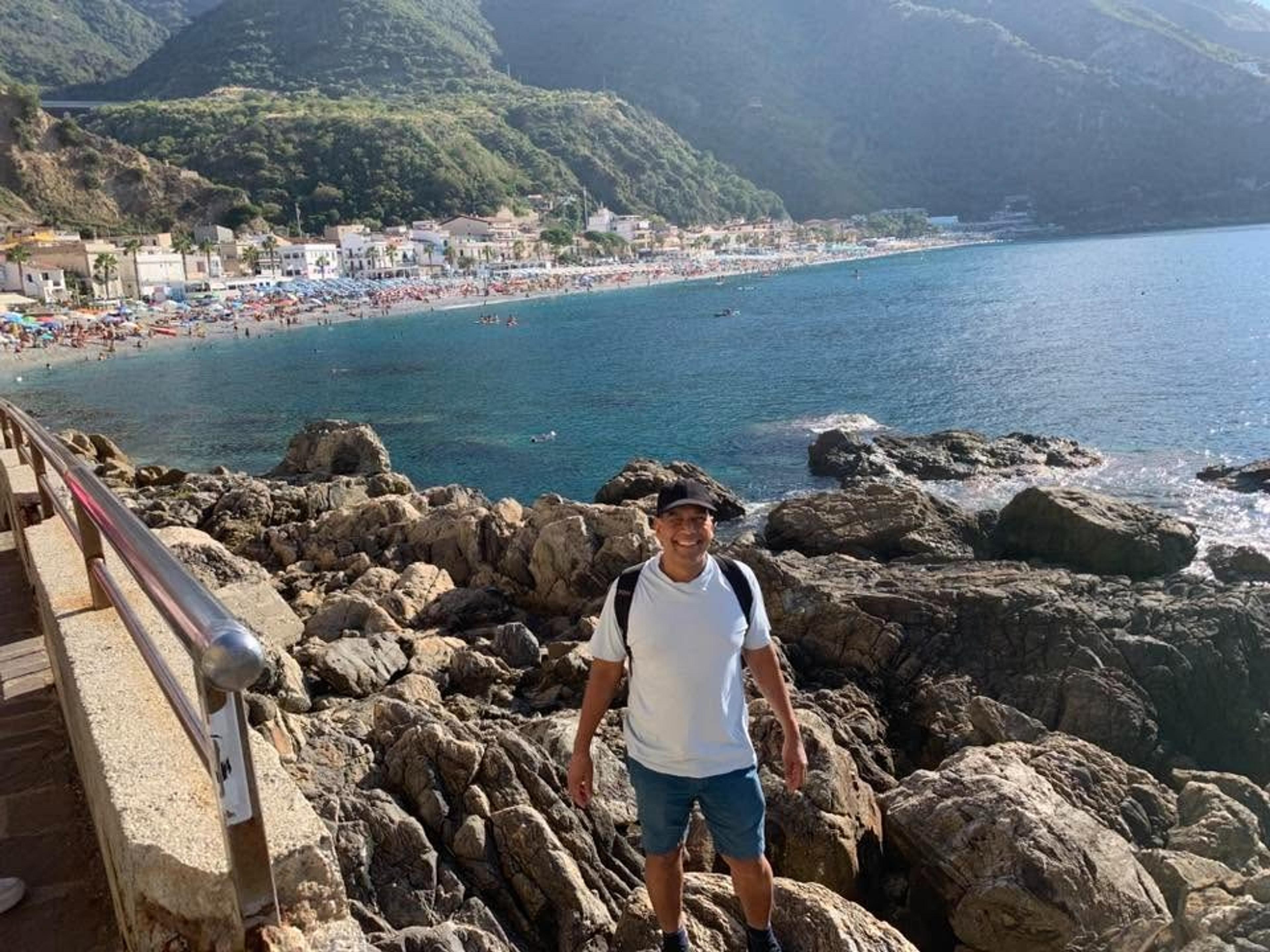 A man stands on a rocky shore with the bustling beach of Scilla, Italy, and its surrounding hills in the background.