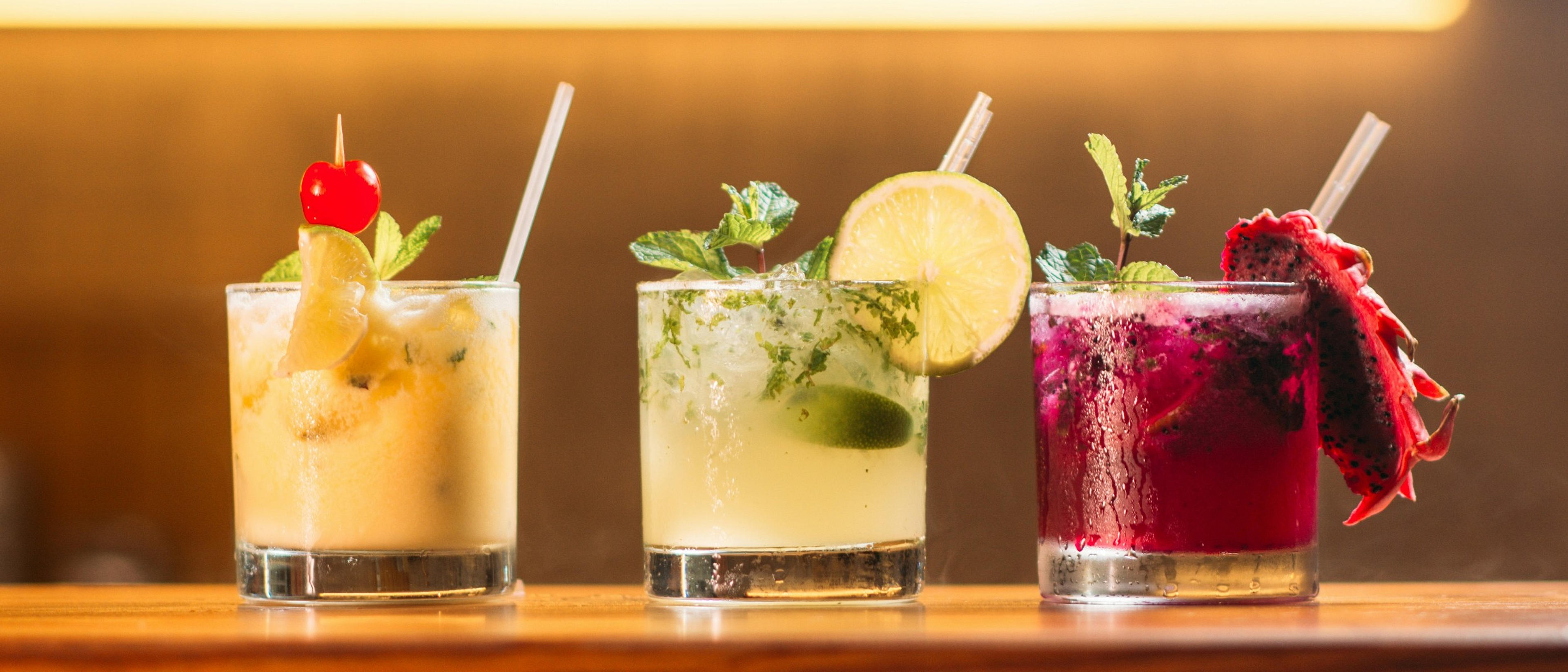 A row of three colorful cocktails garnished with fresh fruit and mint sits on a wooden bar, likely in a cozy lounge.