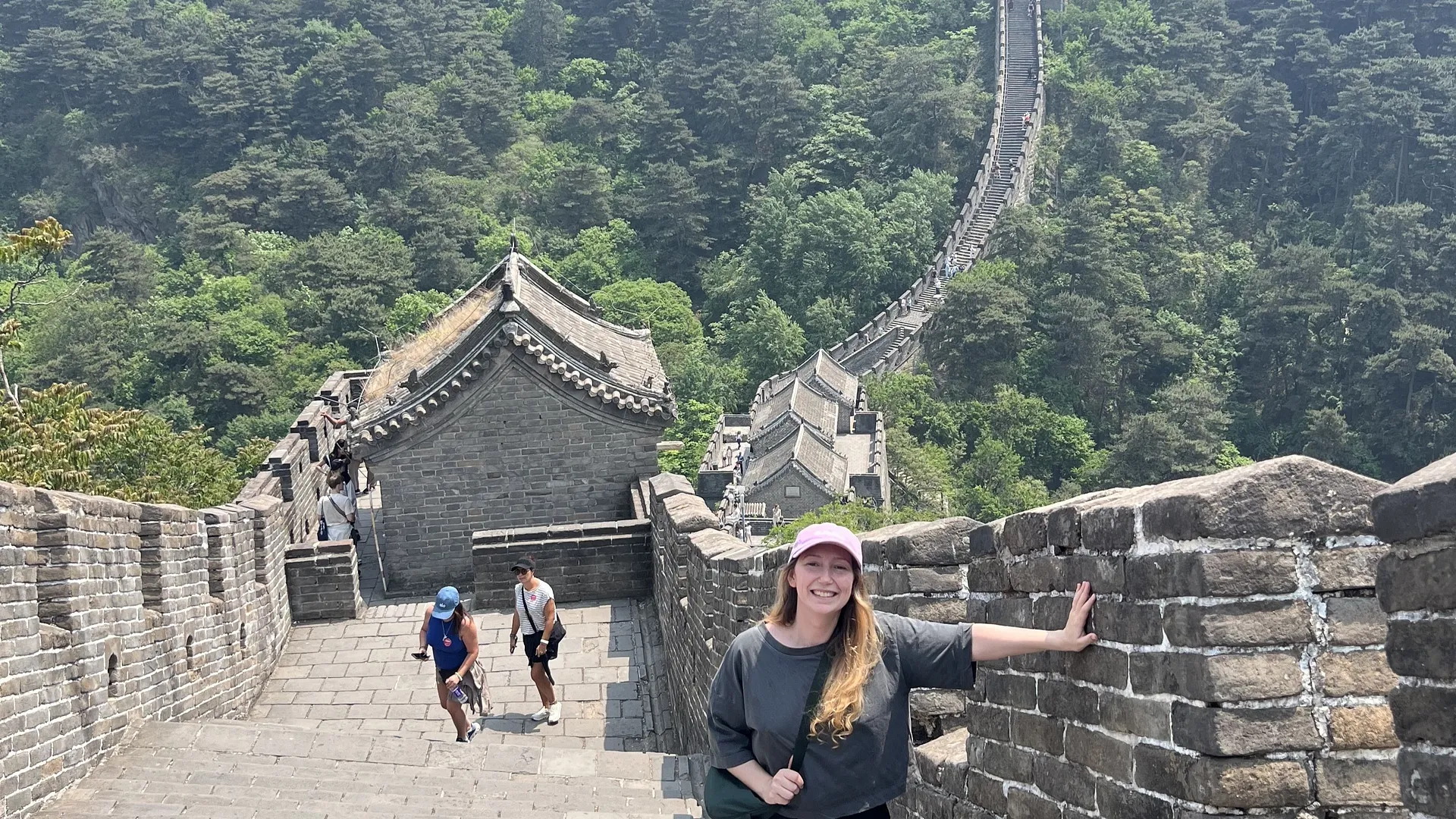 A woman smiles while standing on the Great Wall of China, with lush greenery and a winding section of the wall stretching into the distance.