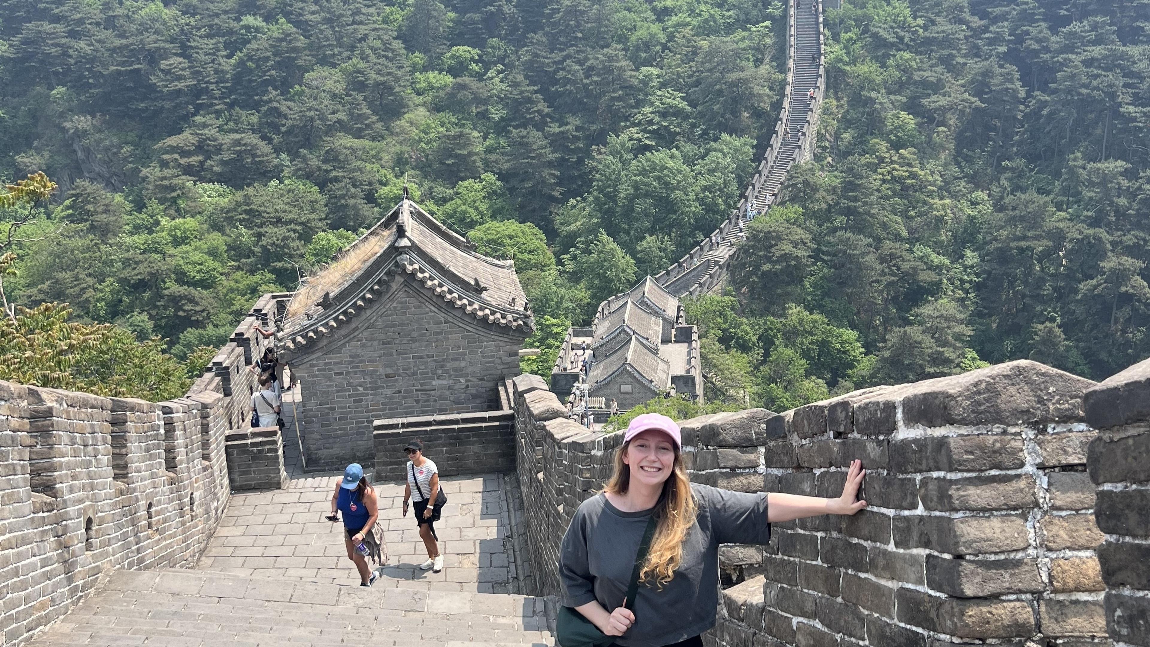 A woman smiles while standing on the Great Wall of China, with lush greenery and a winding section of the wall stretching into the distance.