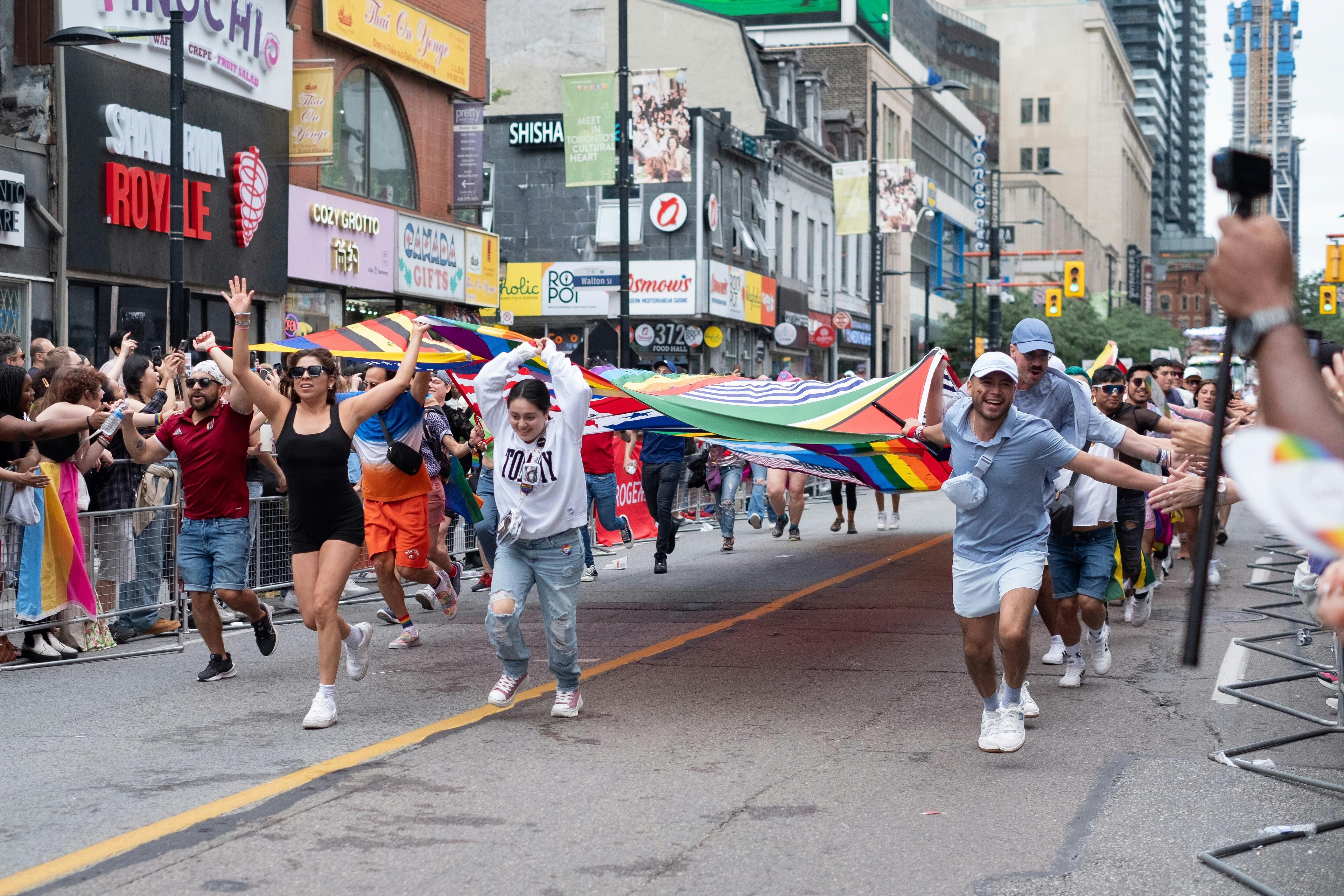 People joyfully run down Church Street in Toronto waving a large rainbow flag during a parade.