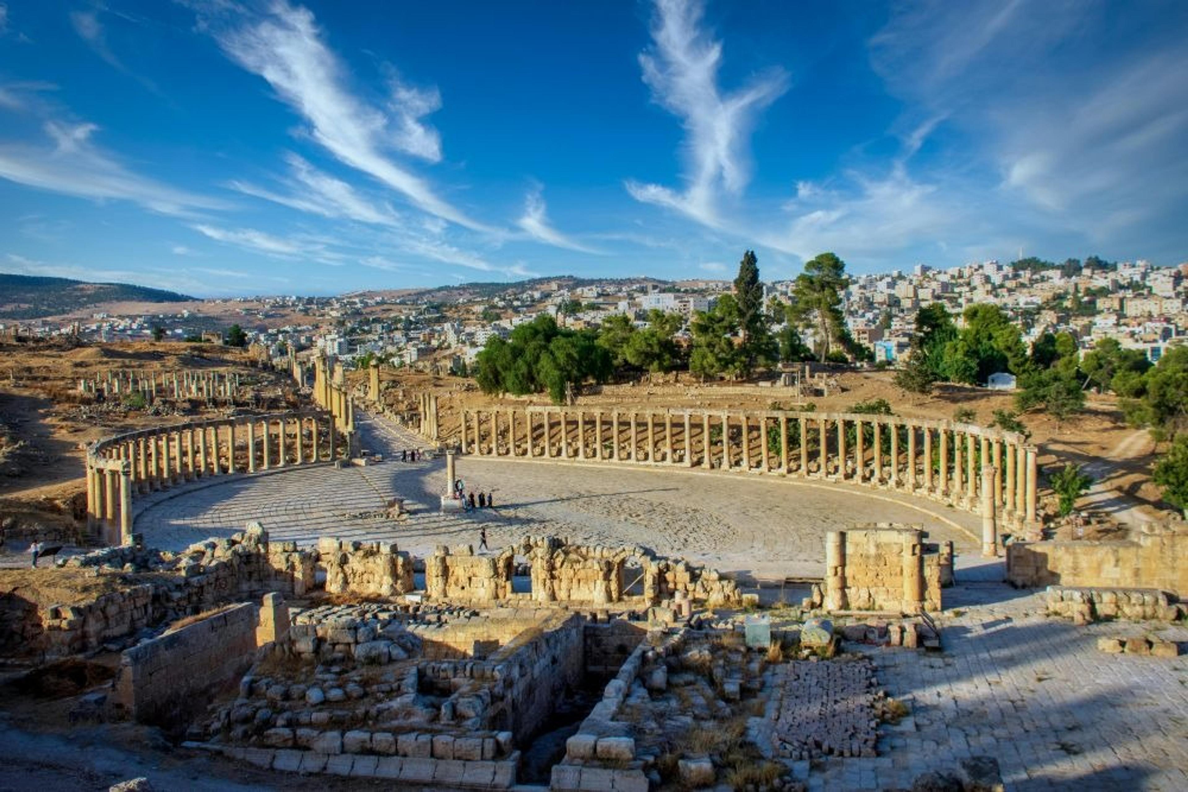 Ancient Roman ruins and the Oval Plaza of Jerash in Jordan stand against a backdrop of arid hills and a sprawling modern town.