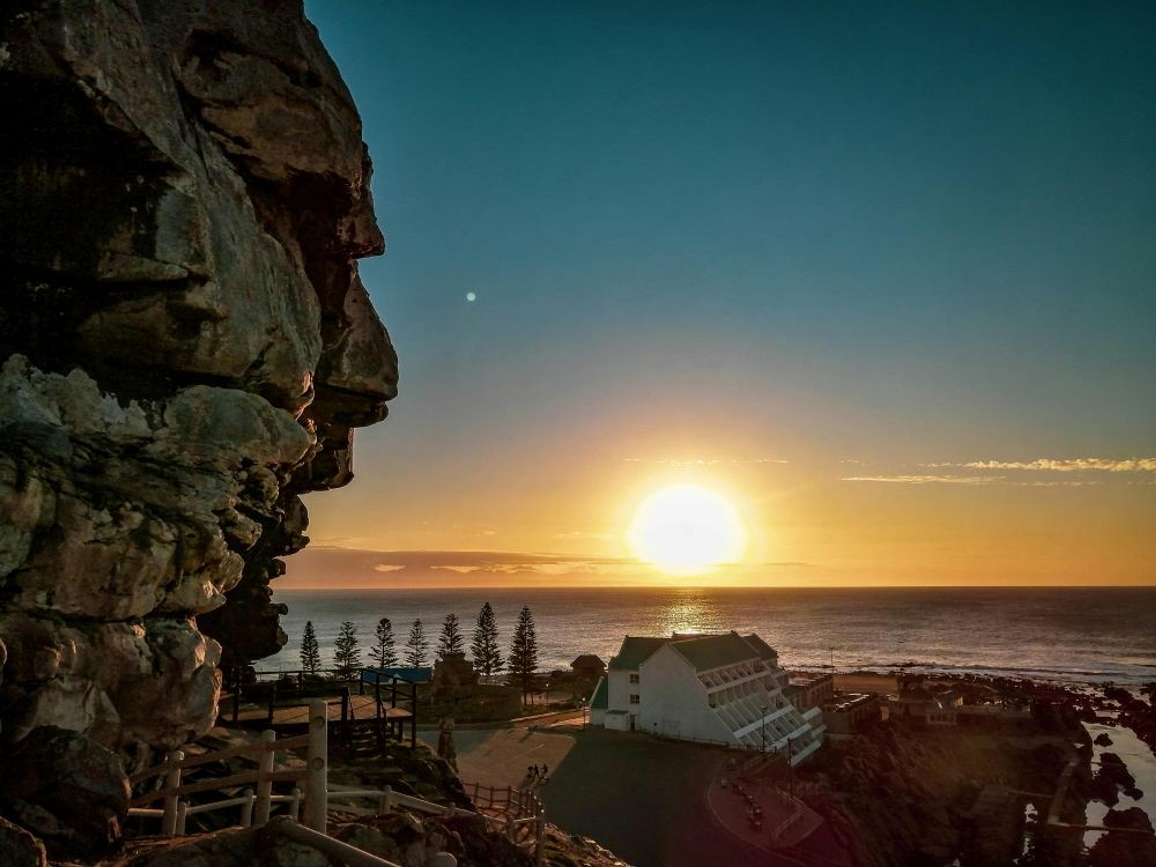 A stunning sunset over the ocean near Mossel Bay, South Africa, with rugged cliffs and a coastal building in view.