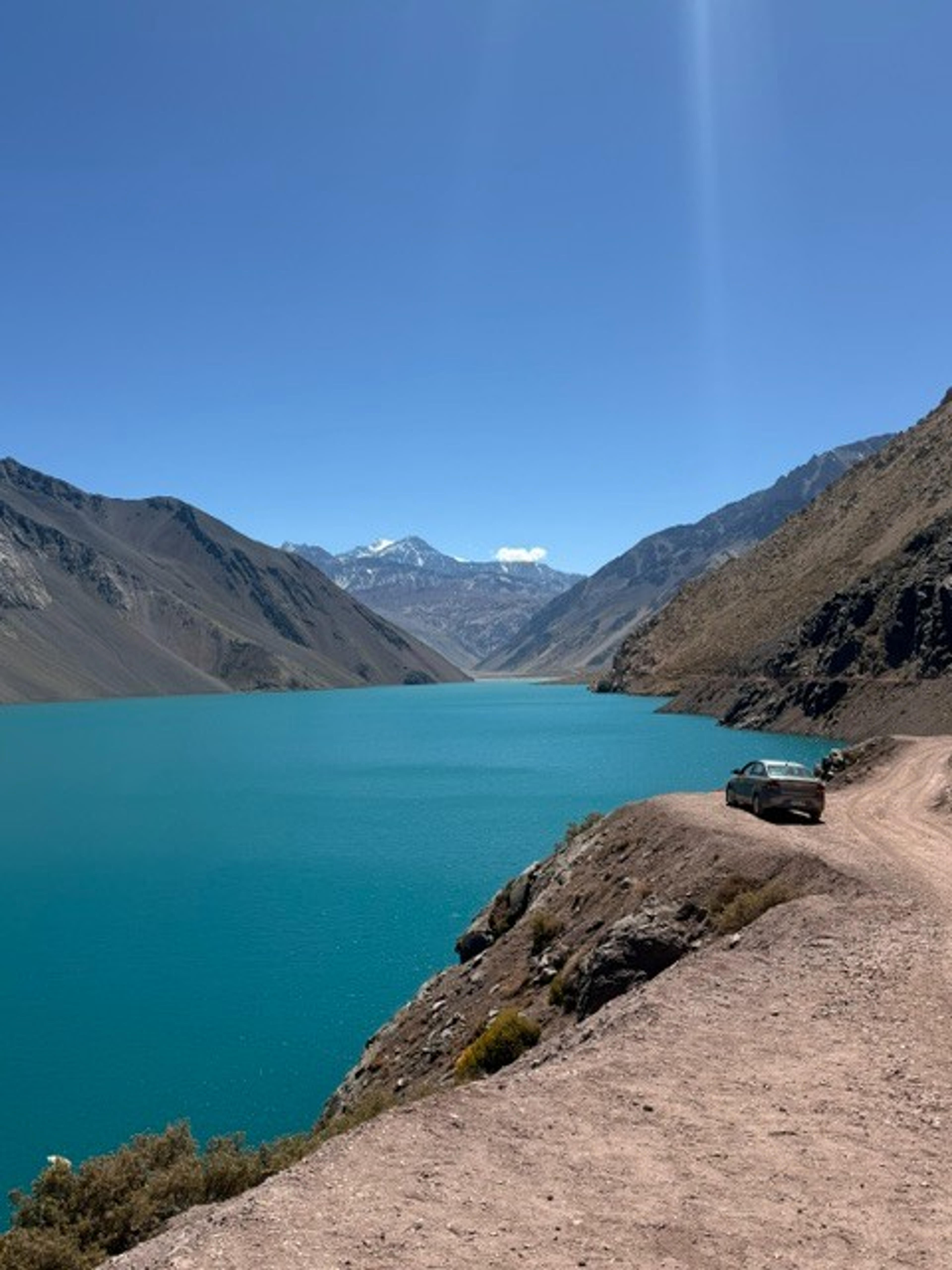 A turquoise lake is surrounded by rugged mountains and a dirt road in Cajón del Maipo, Chile.