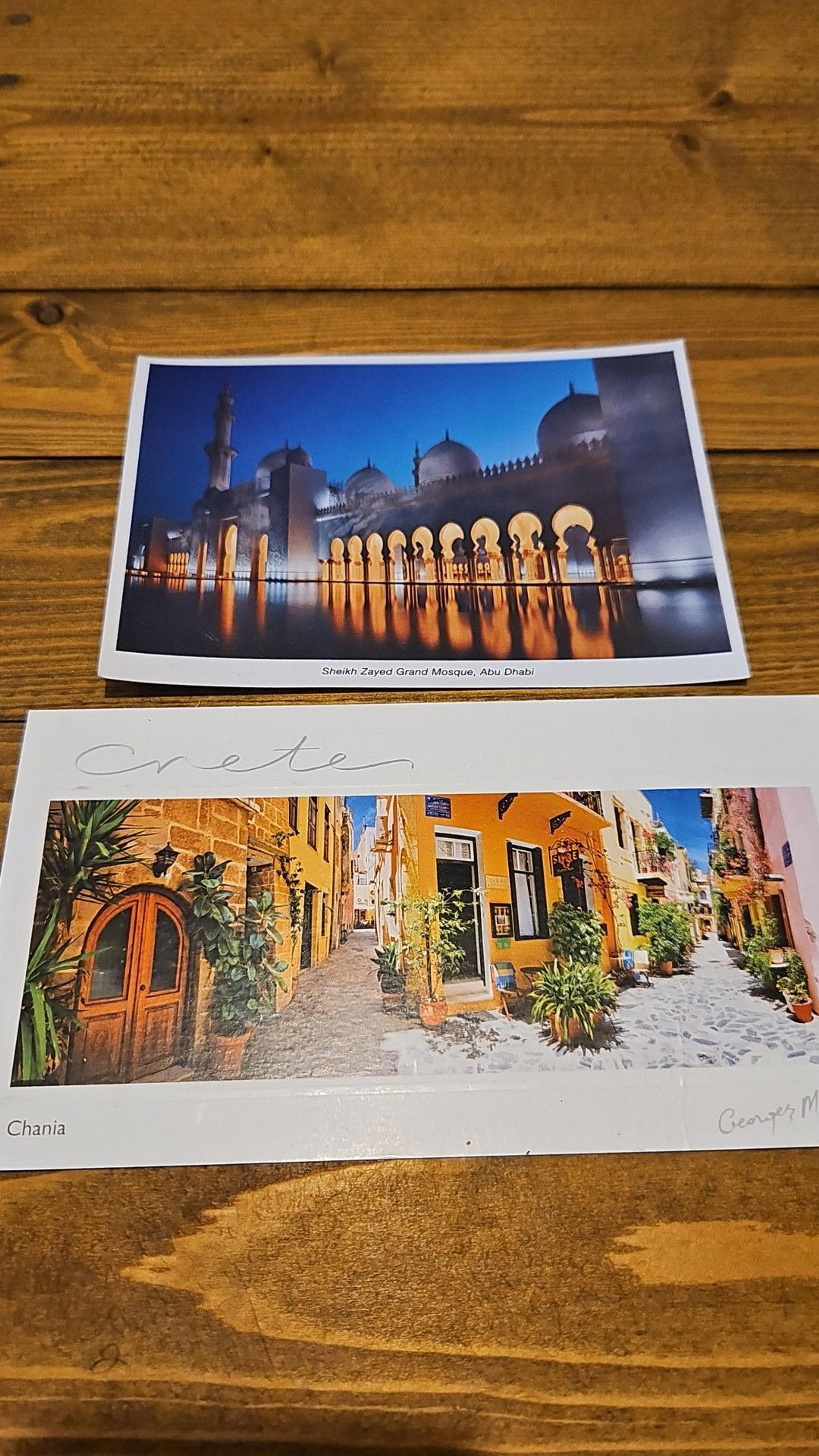 Two postcards are placed on a wooden table, depicting Sheikh Zayed Grand Mosque in Abu Dhabi and a charming street in Chania, Crete.
