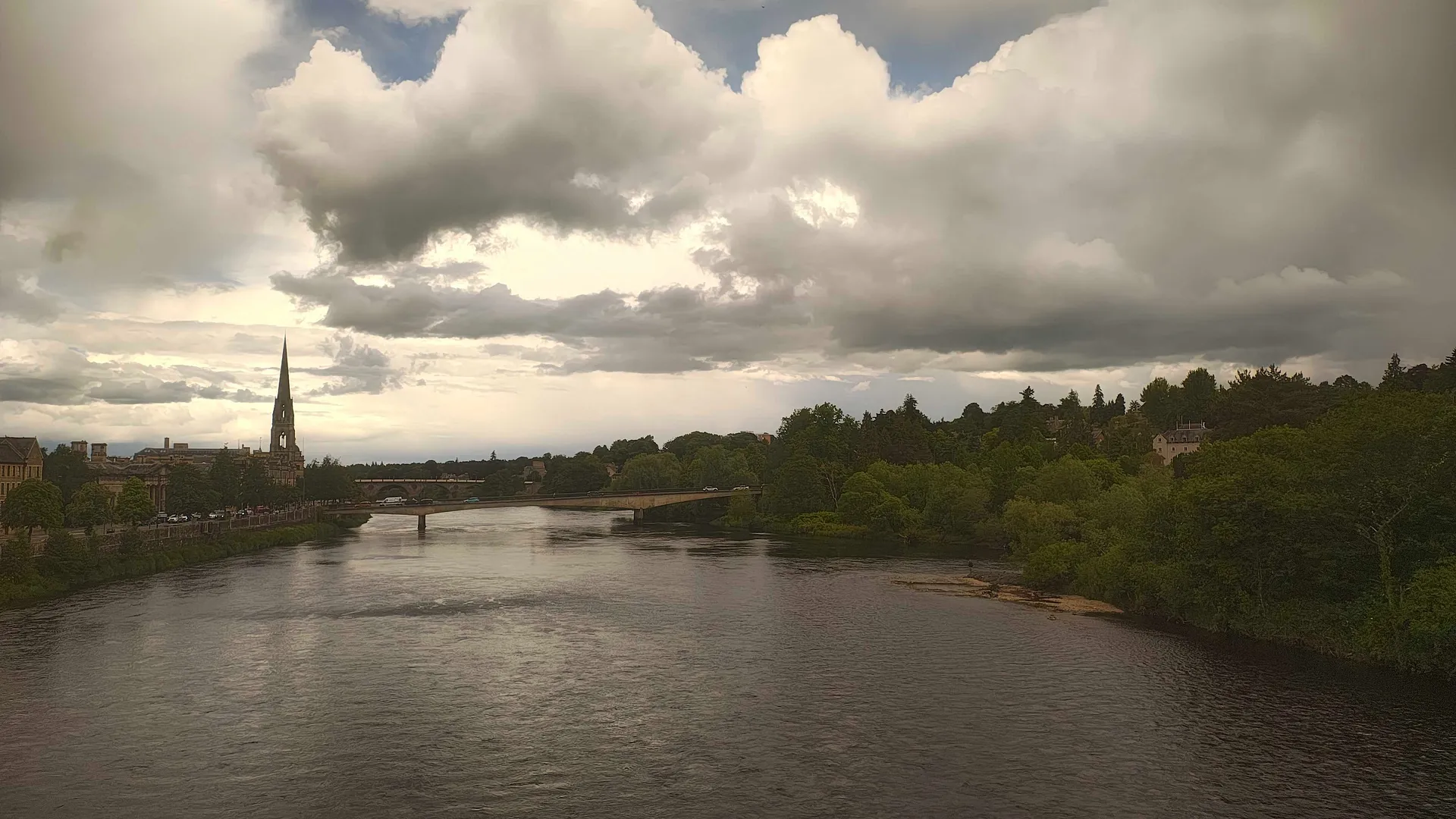 Perth, Scotland's riverside view features a dramatic cloudy sky, lush greenery, and a distinctive church spire.