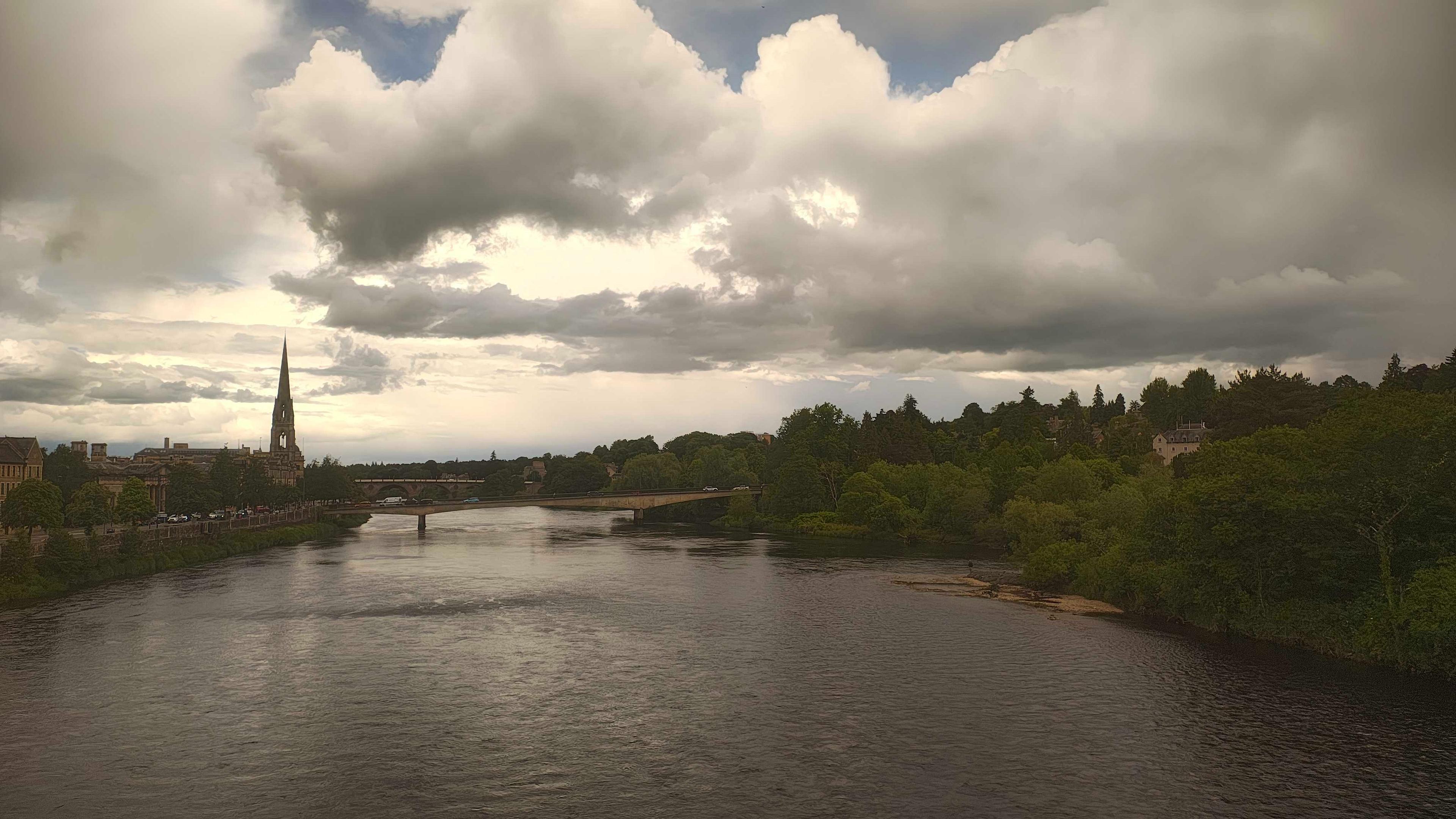 Perth, Scotland's riverside view features a dramatic cloudy sky, lush greenery, and a distinctive church spire.
