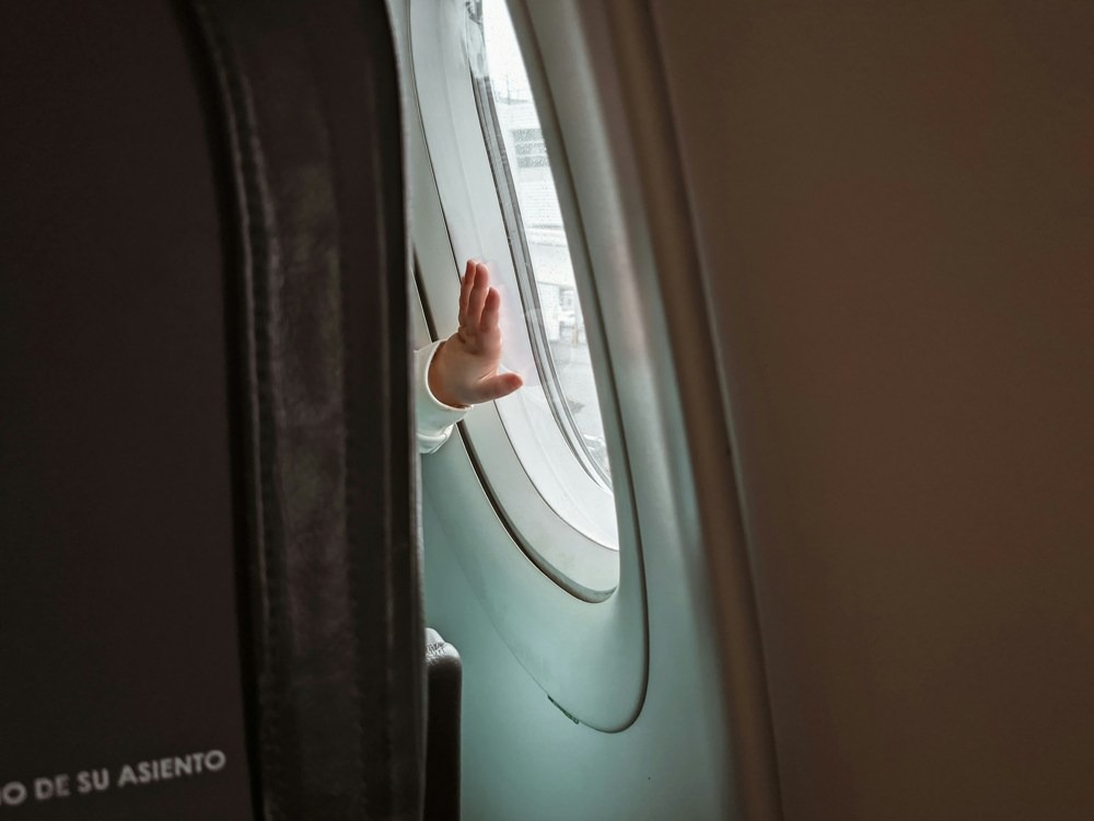 A child's hand waves from an airplane window, with the text "DE SU ASIENTO" visible on the seat.
