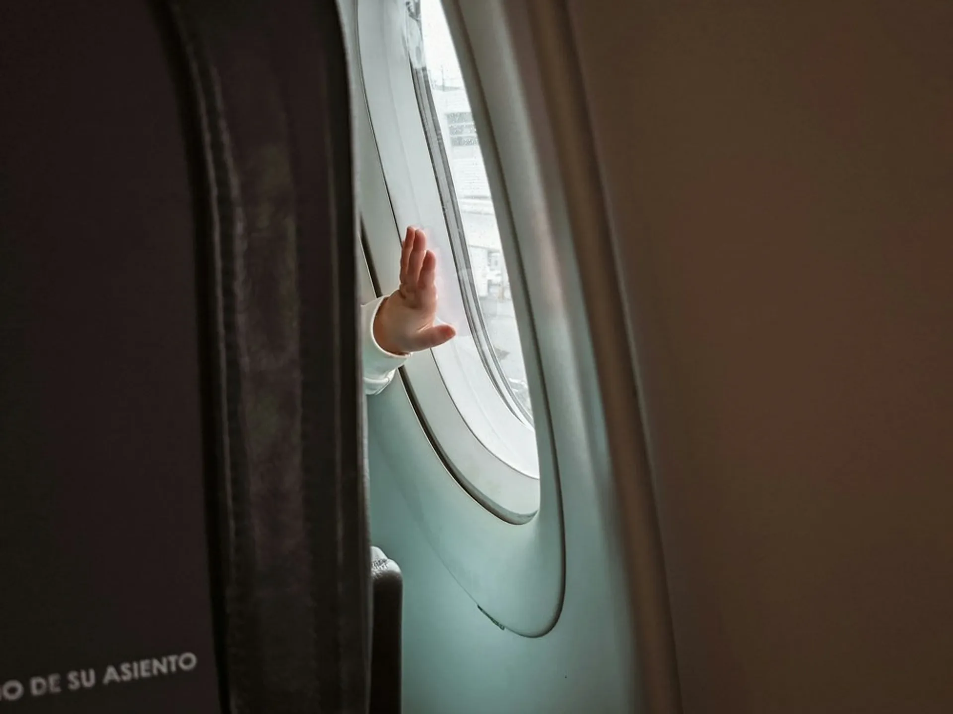 A child's hand waves from an airplane window, with the text "DE SU ASIENTO" visible on the seat.