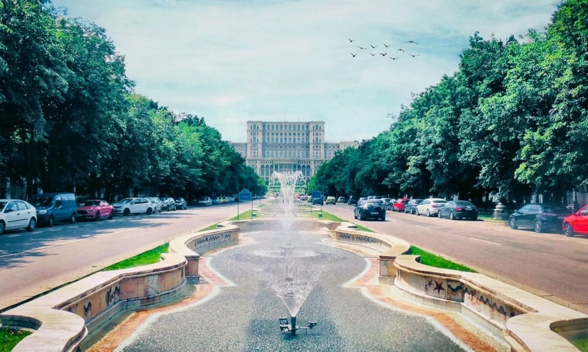 Fountains line a boulevard leading to the massive Palace of the Parliament in Bucharest, Romania, with lush trees bordering the road.