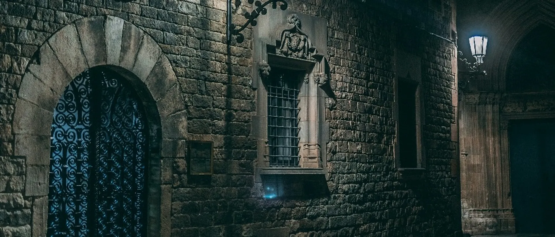 An atmospheric nighttime view captures the historic Gothic Quarter in Barcelona, featuring a stone facade with intricate ironwork and a lantern casting a warm glow.