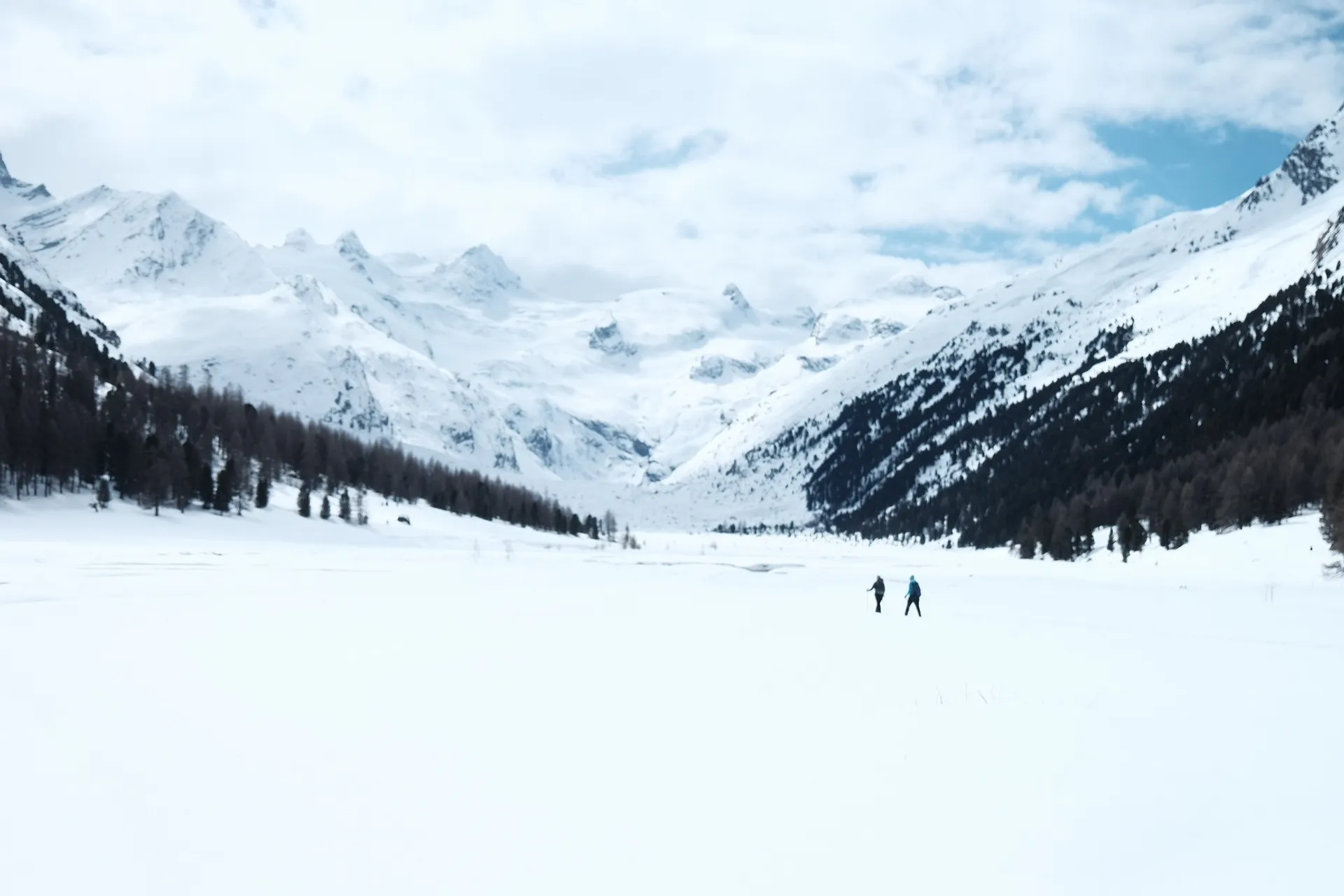 Two people traverse a snow-covered valley surrounded by the majestic Alps in Switzerland.