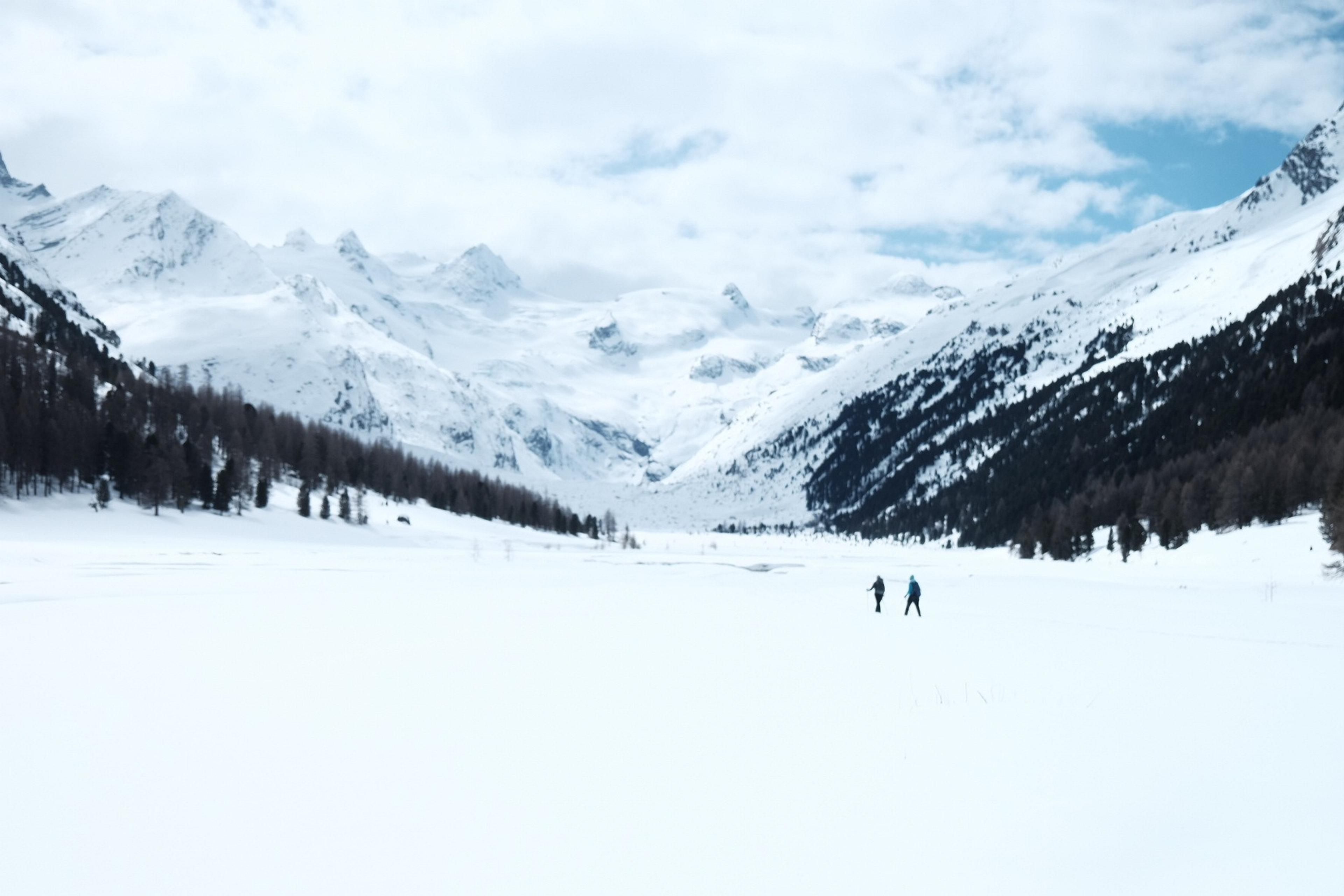 Two people traverse a snow-covered valley surrounded by the majestic Alps in Switzerland.