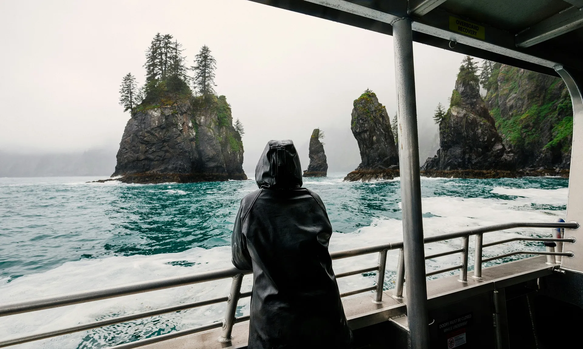 A person in a hooded raincoat gazes at the rocky, tree-topped sea stacks of Kenai Fjords in Alaska from the deck of a boat.