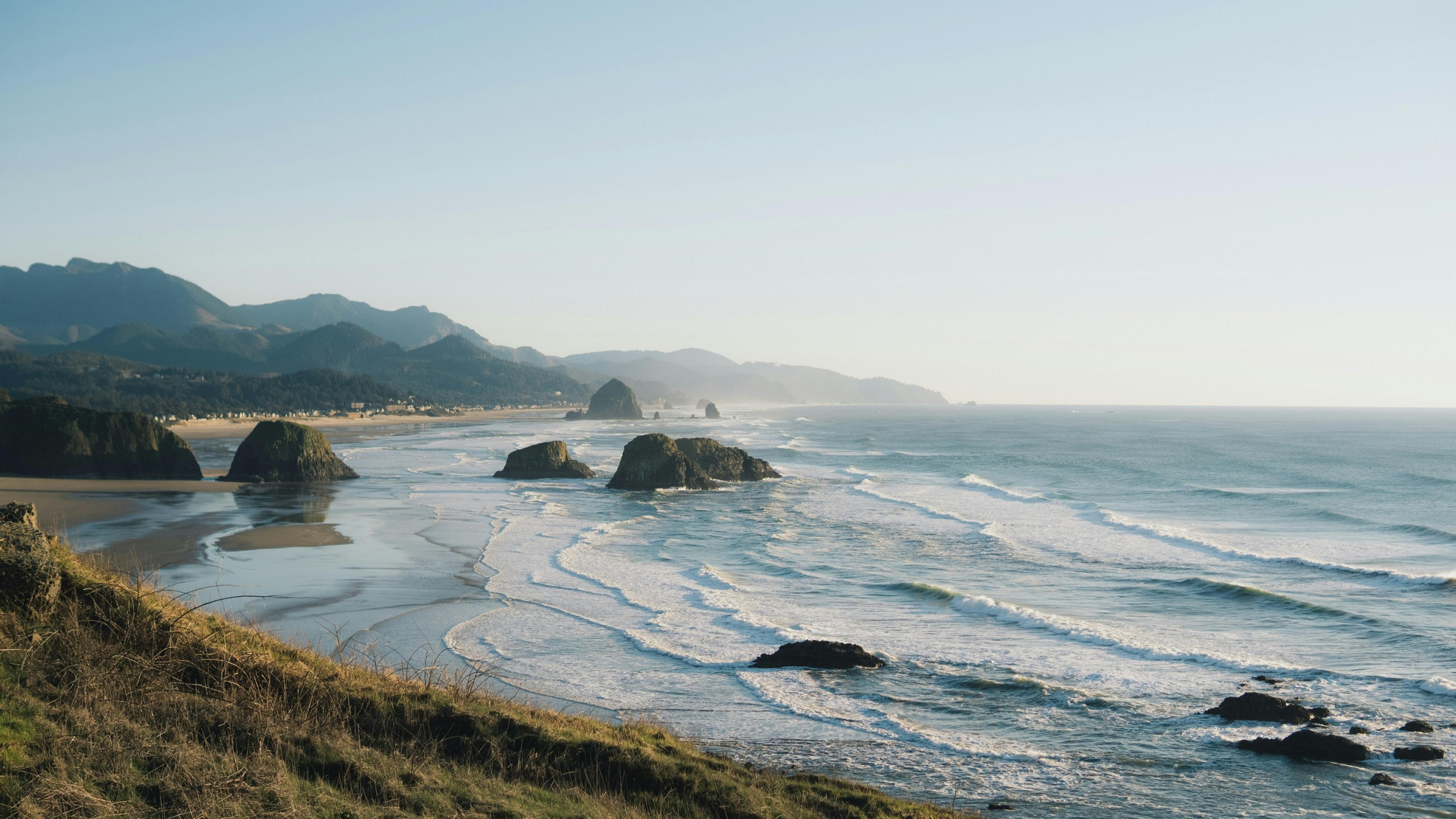 Waves gently crash against the iconic sea stacks of Cannon Beach, Oregon, with hazy mountains in the background.