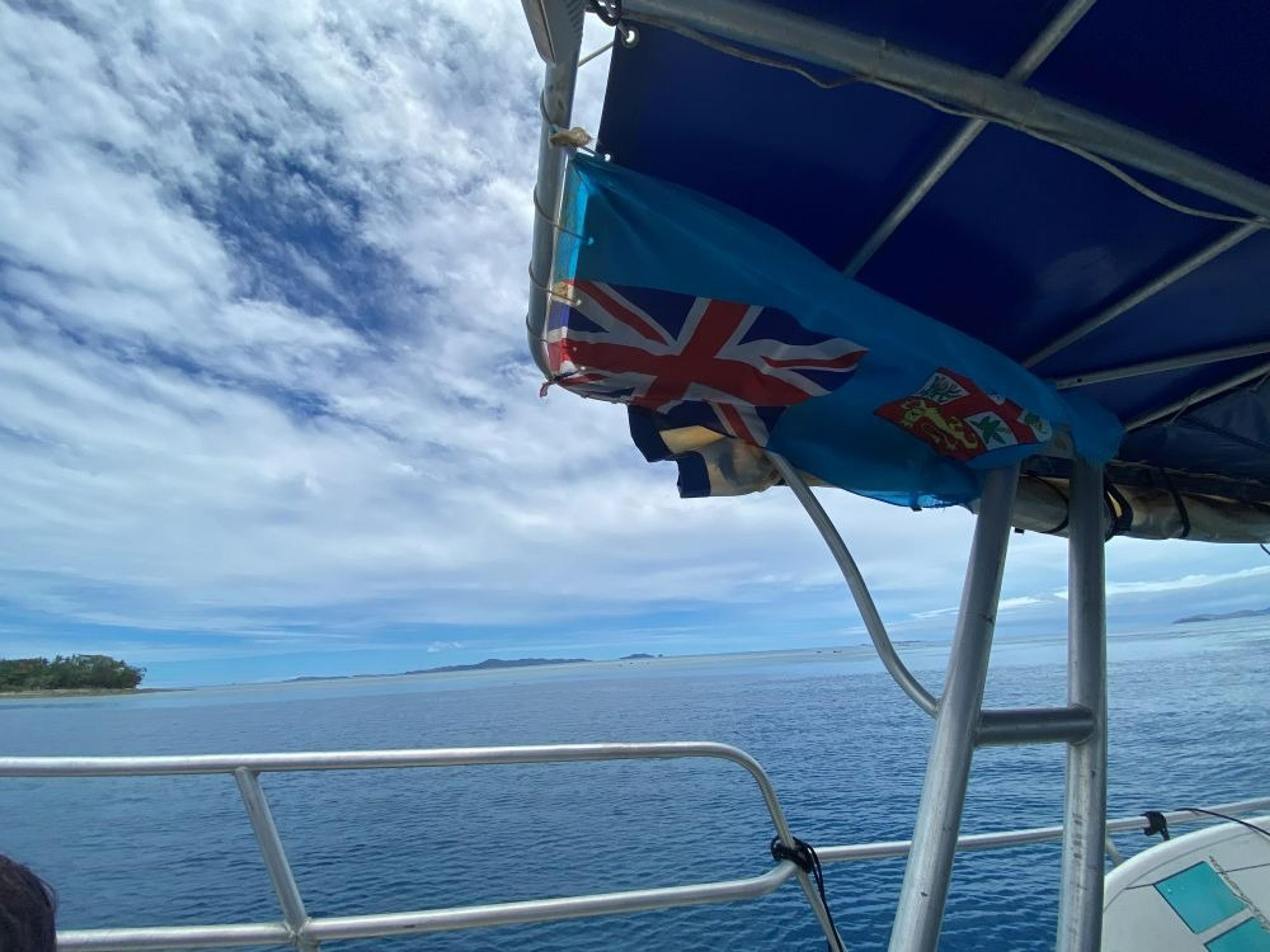 A boat sails near the islands of Fiji, displaying the national flag against a backdrop of clear blue skies and calm waters.