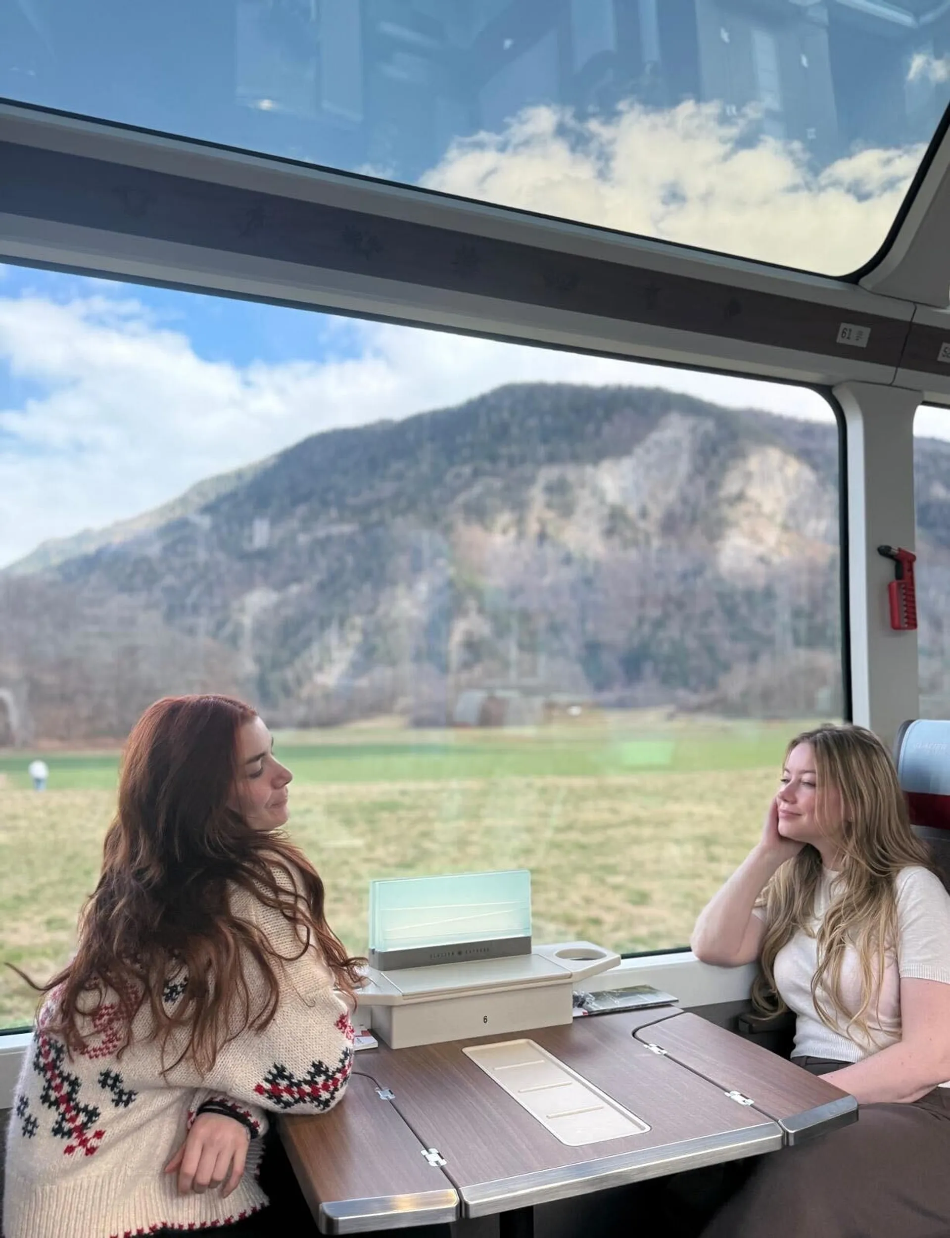 Two people enjoy a scenic train ride through the Swiss Alps, with mountains visible through large panoramic windows.