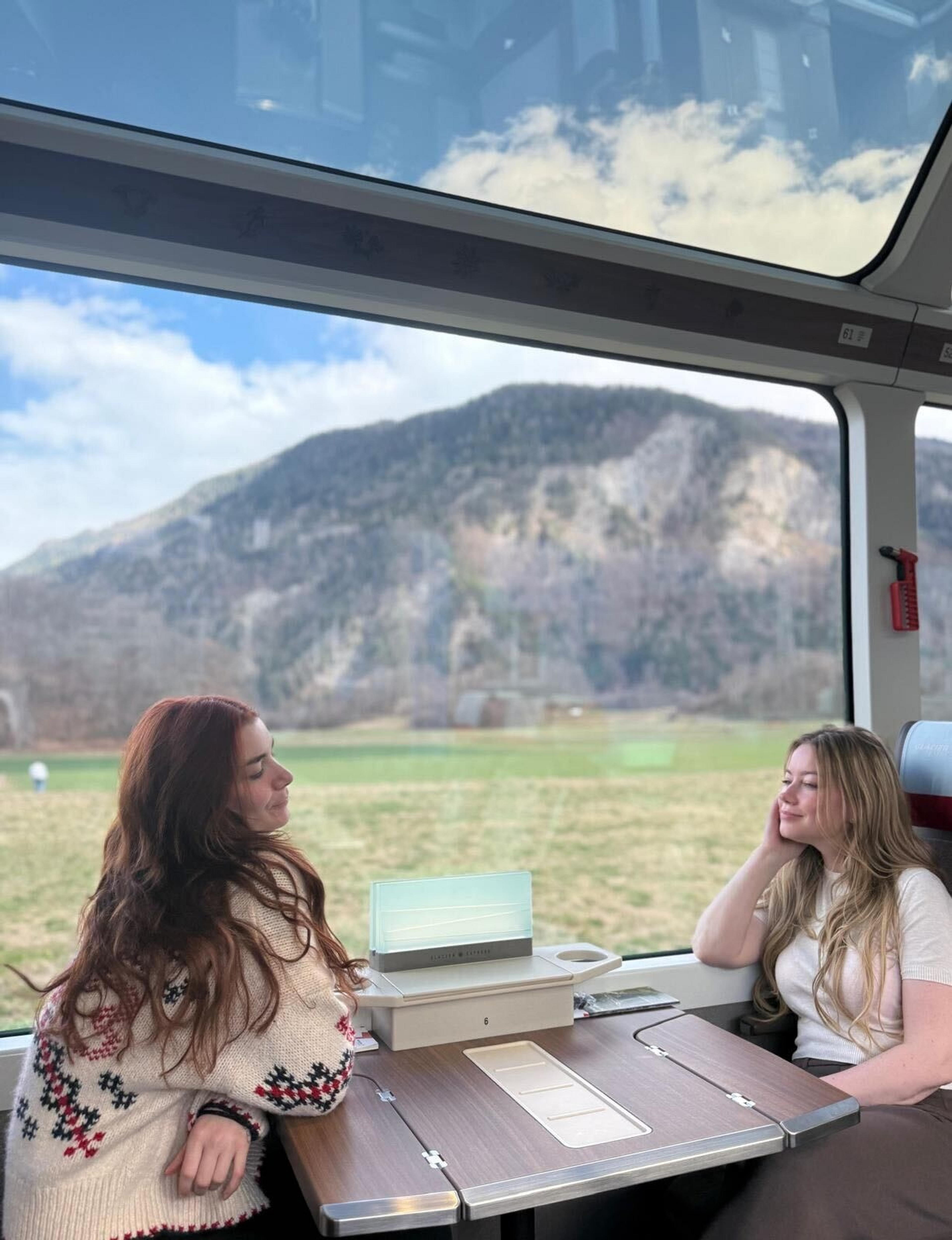 Two people enjoy a scenic train ride through the Swiss Alps, with mountains visible through large panoramic windows.