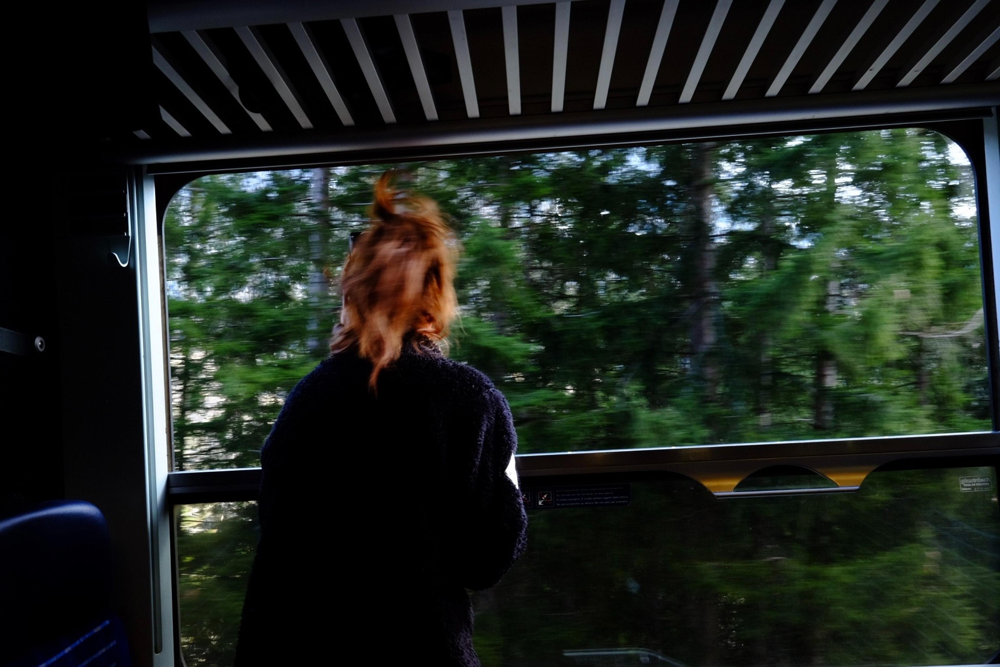A person with red hair gazes out the window of a train as lush green trees blur past.