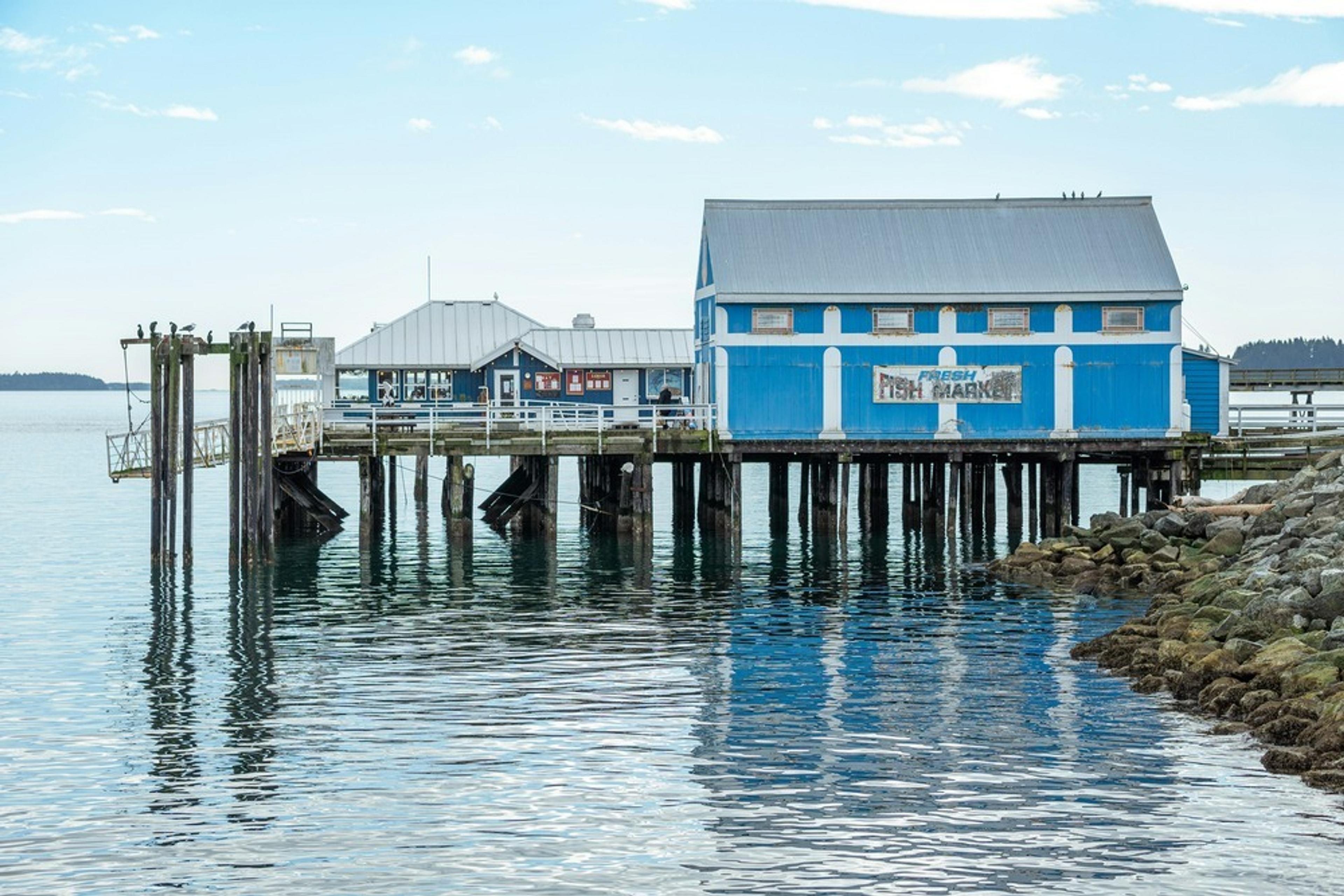 A picturesque wharf in Sidney, British Columbia, featuring a blue building serving as a fresh fish market on stilts above calm waters.