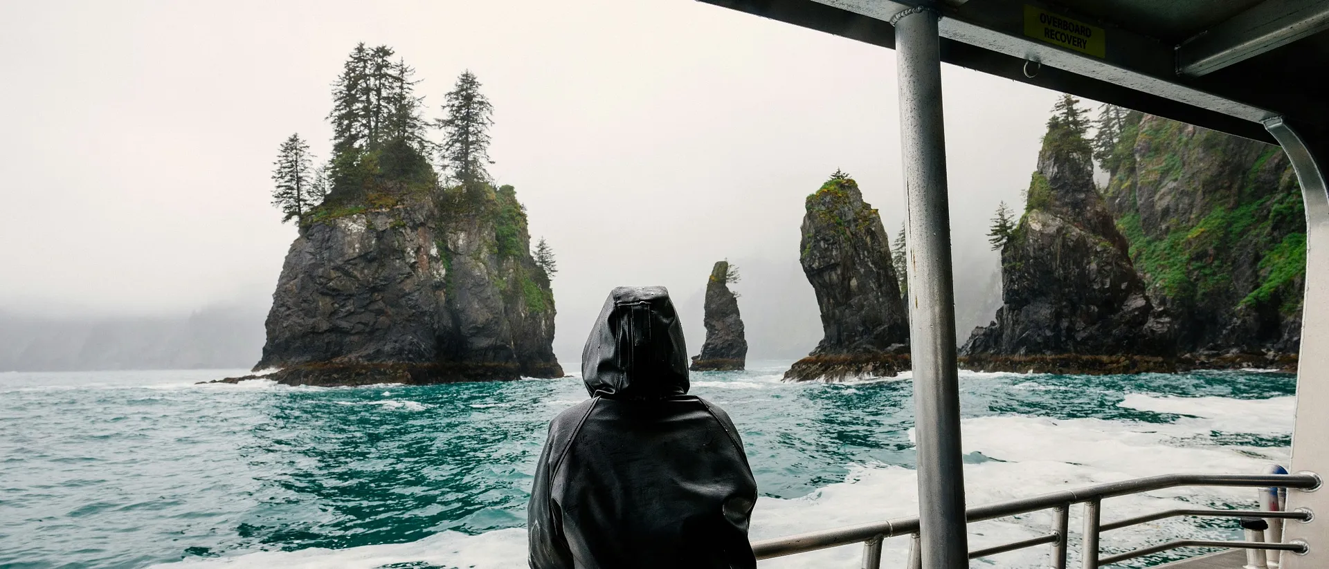 A person in a raincoat gazes at the rugged sea stacks and misty cliffs of Kenai Fjords National Park, Alaska, from a boat.