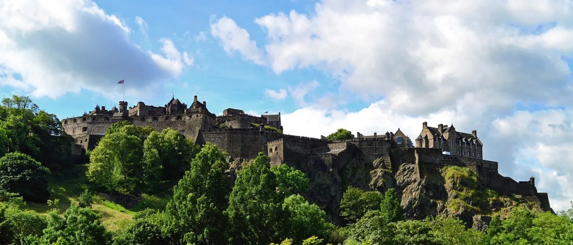 Edinburgh Castle sits majestically atop Castle Rock, surrounded by lush greenery under a partly cloudy sky.