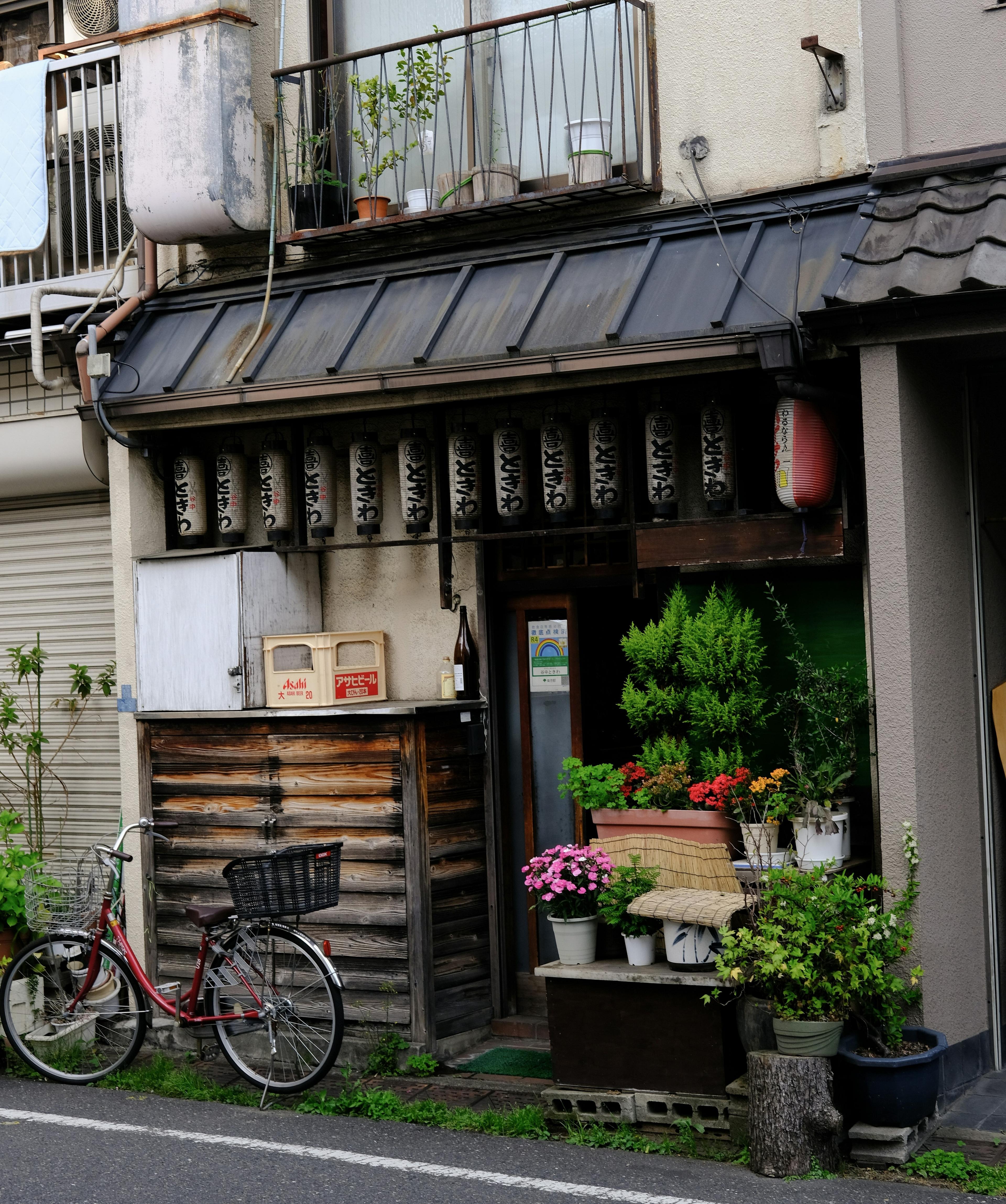 A quaint street in Japan features a small traditional shop adorned with hanging lanterns, potted plants, and a parked bicycle.