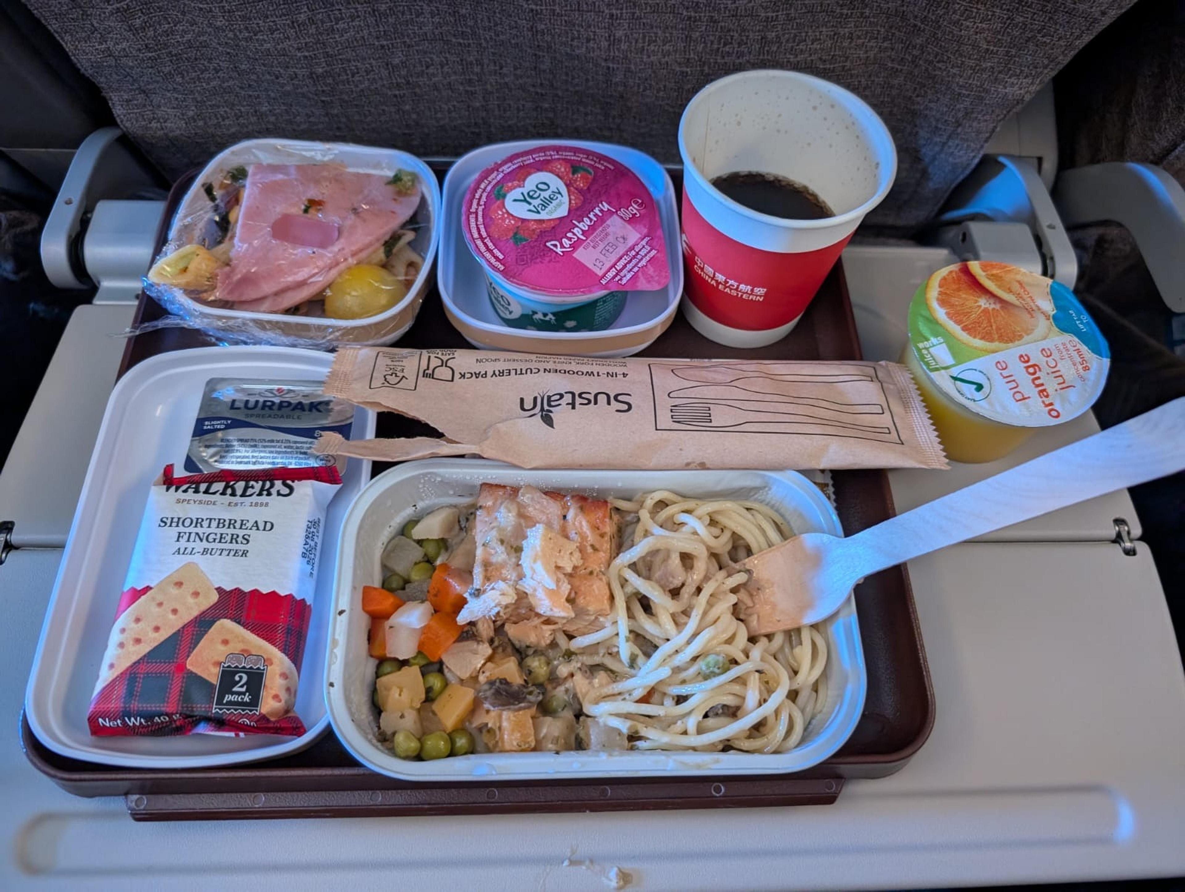 A meal tray on a China Eastern Airlines flight features salmon spaghetti with vegetables, ham and potato salad, raspberry yogurt, orange juice, and shortbread cookies.