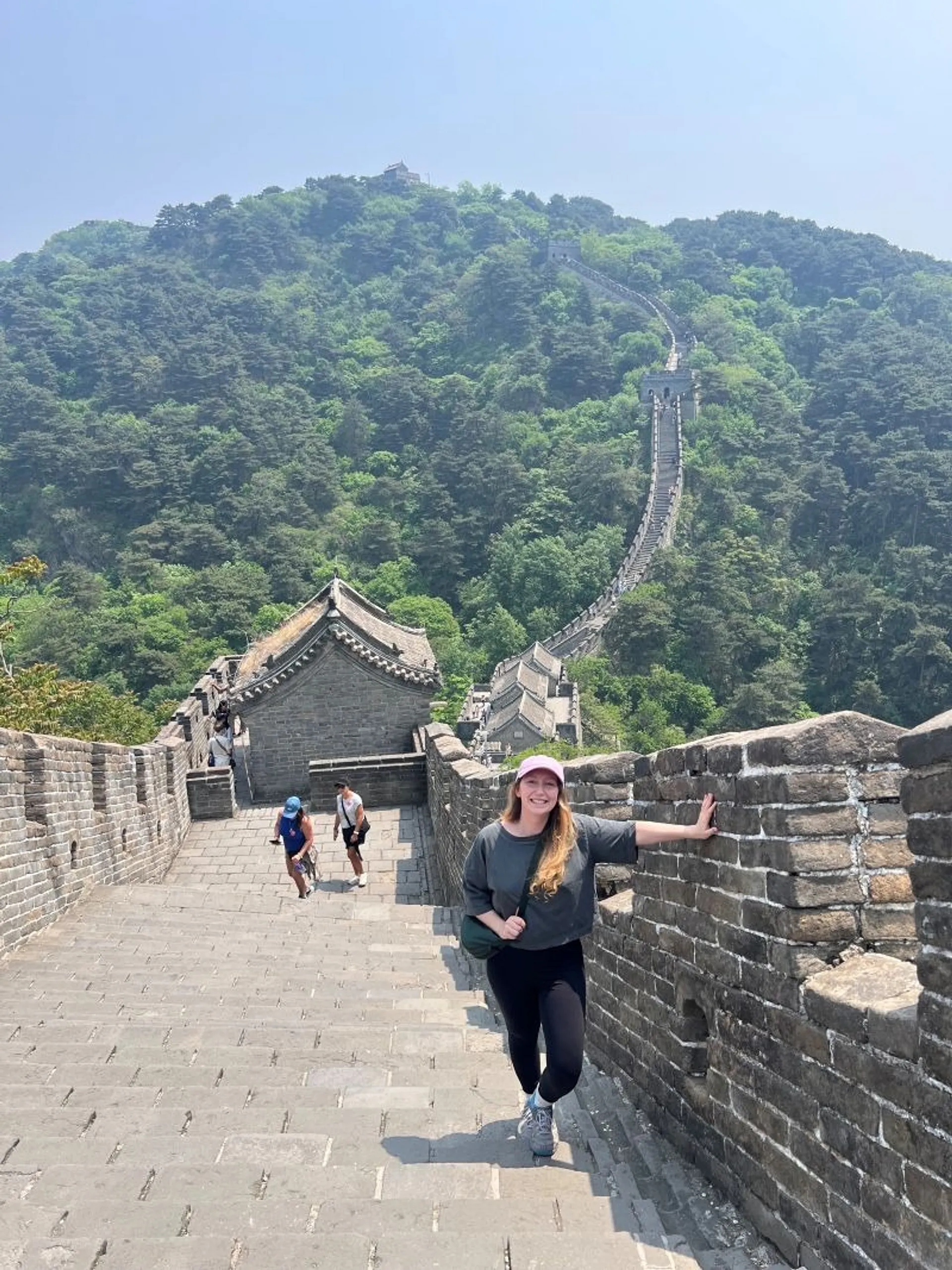 A woman smiles while walking along the Great Wall of China amidst lush, green hills.