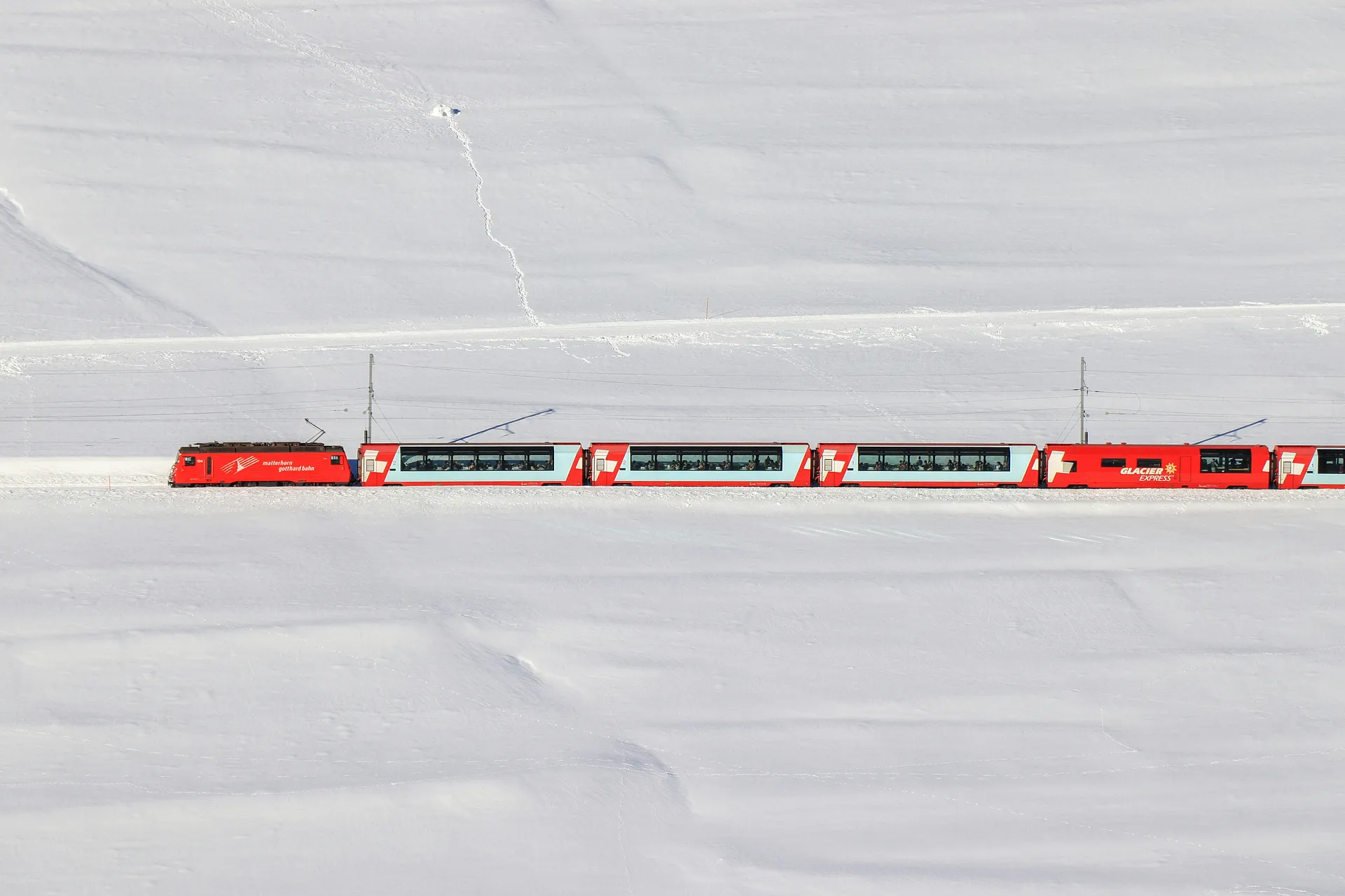 A bright red Glacier Express train travels through the snowy landscape of the Swiss Alps.