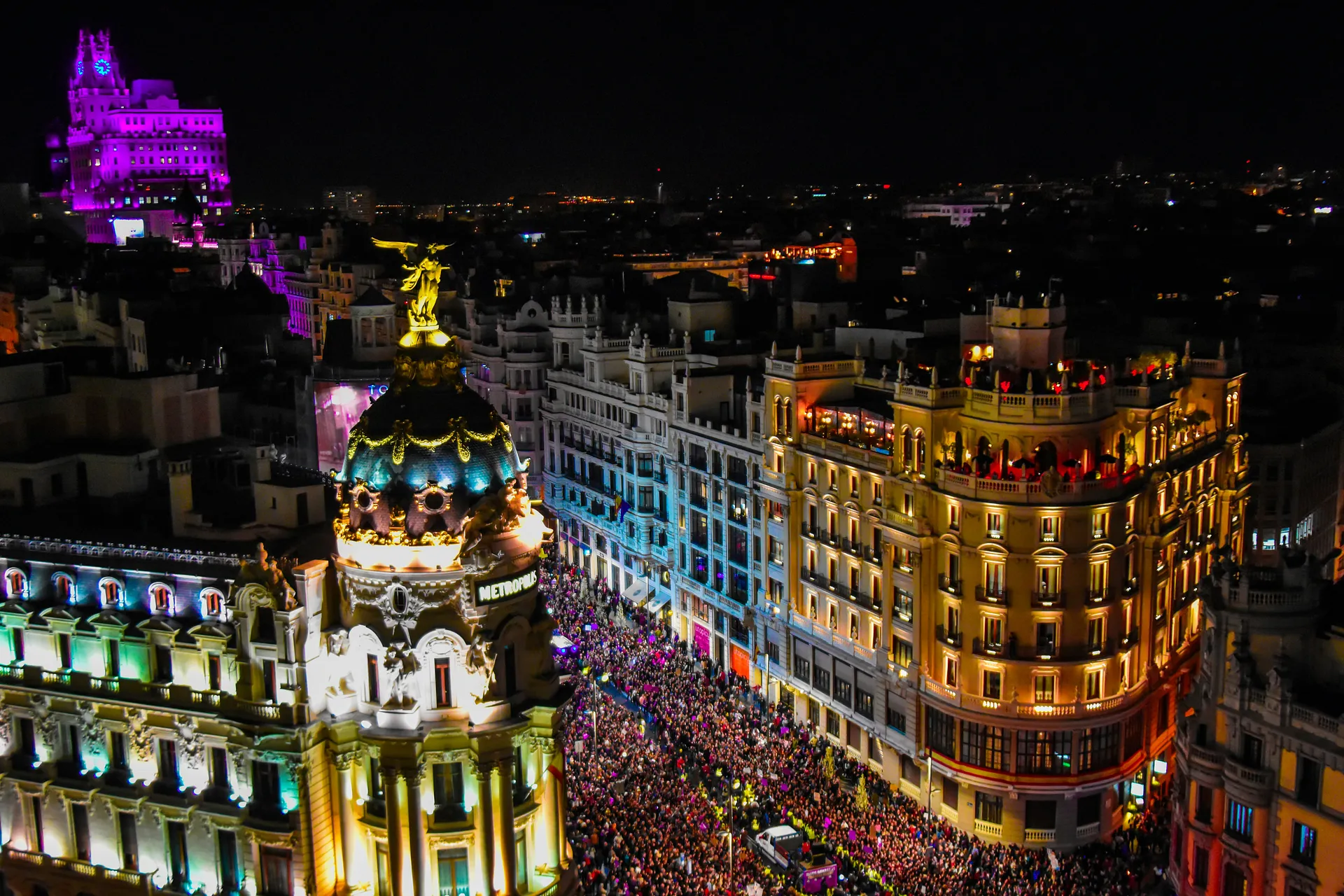 A vibrant nighttime scene captures the bustling Gran Vía in Madrid, with the iconic Metropolis Building illuminated and crowds filling the street.