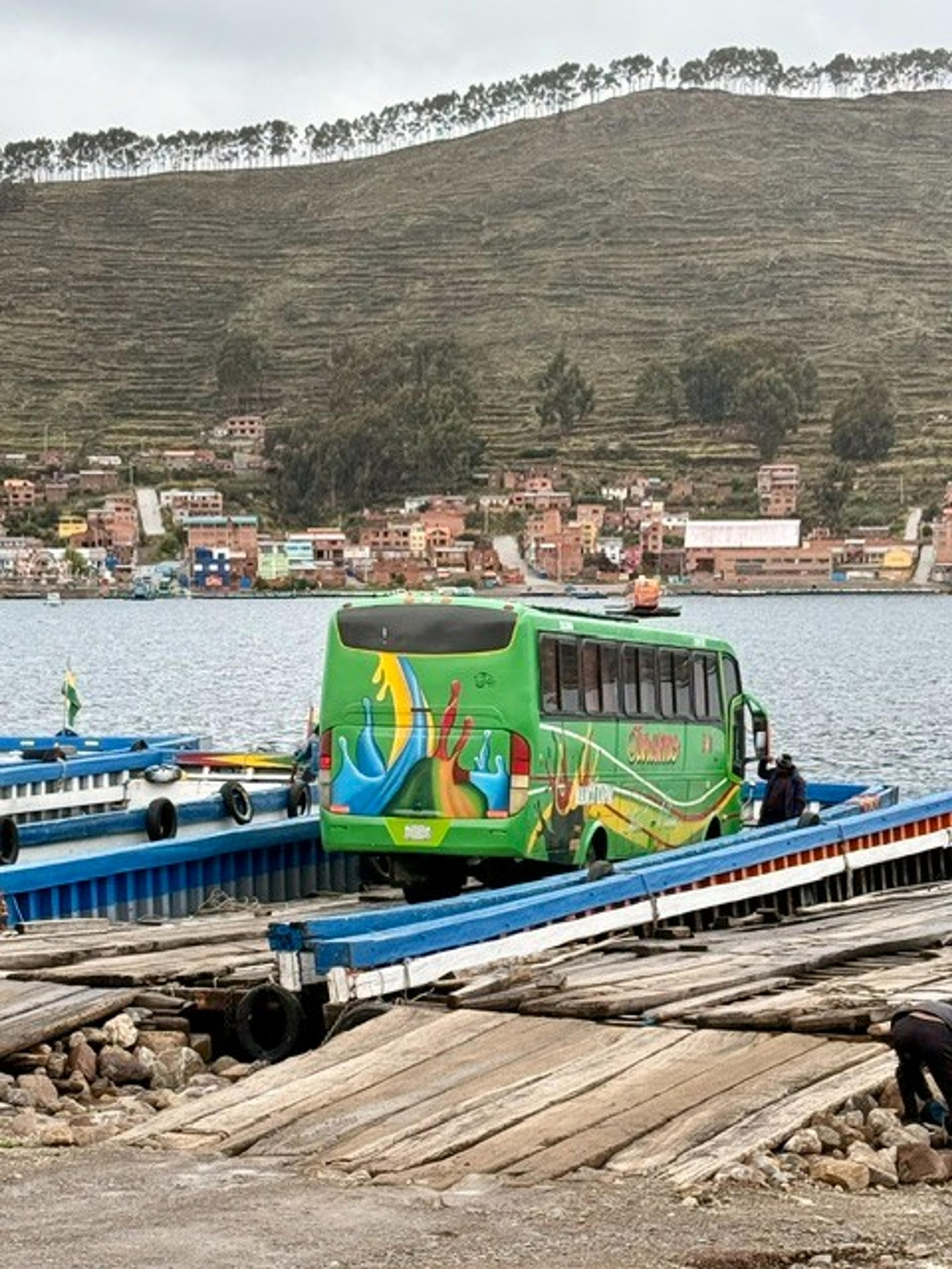 A vibrant green bus is ferried across Lake Titicaca near Copacabana, Bolivia, with a backdrop of hills and colorful village buildings.
