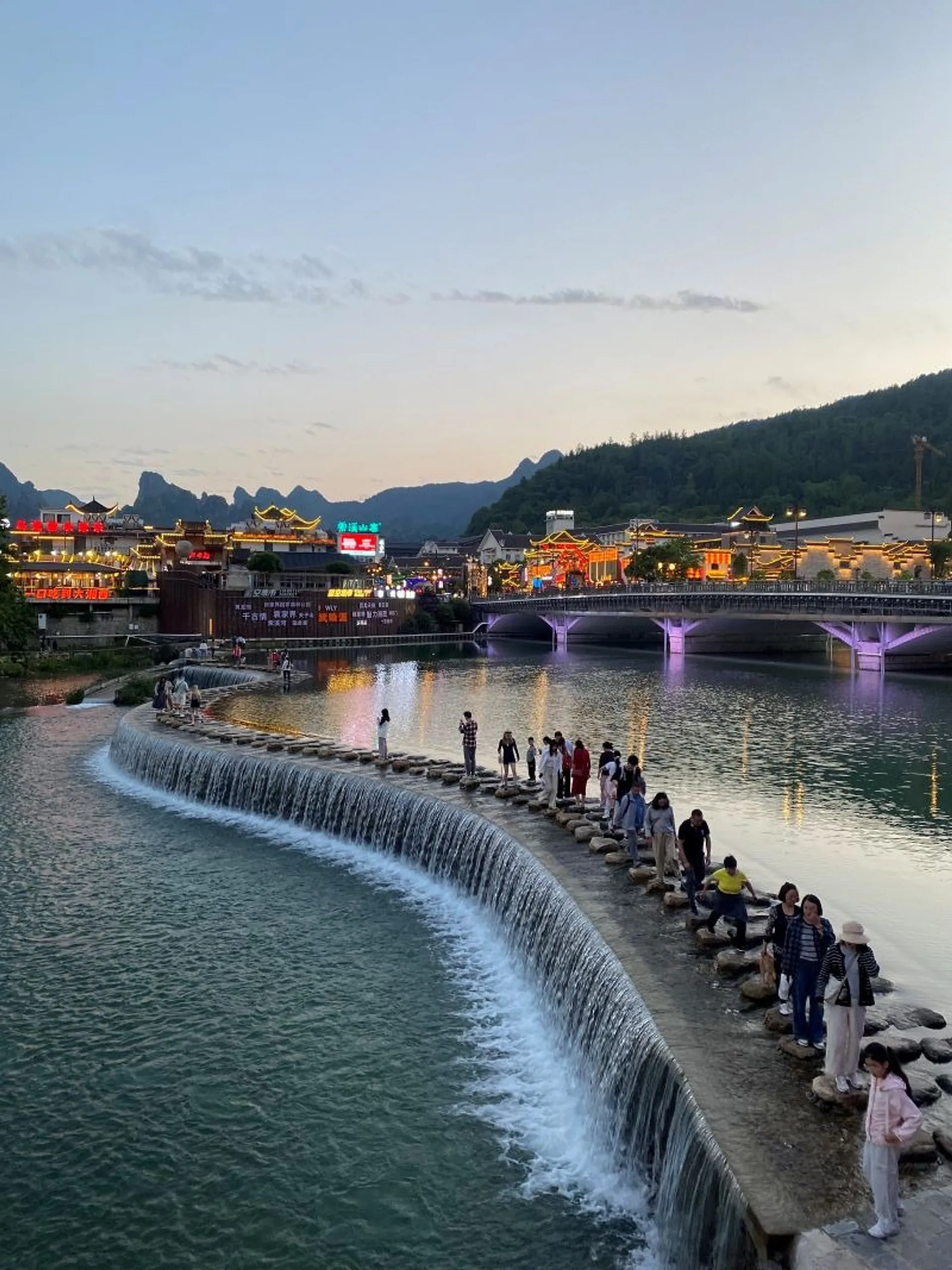 At dusk, people gather on stepping stones across the Xingping Waterfall in Lishui, with illuminated buildings and mountains in the background.