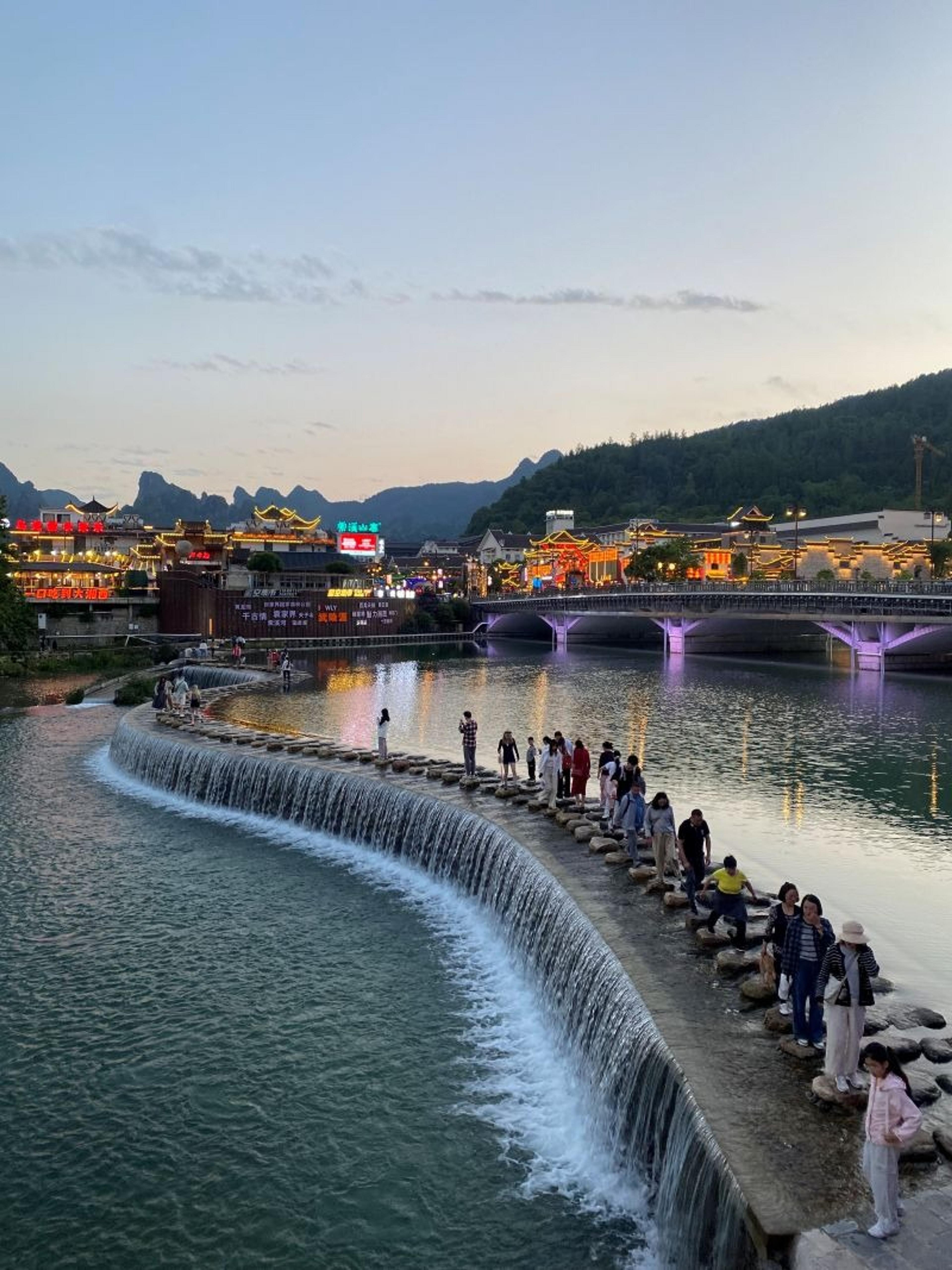 At dusk, people gather on stepping stones across the Xingping Waterfall in Lishui, with illuminated buildings and mountains in the background.