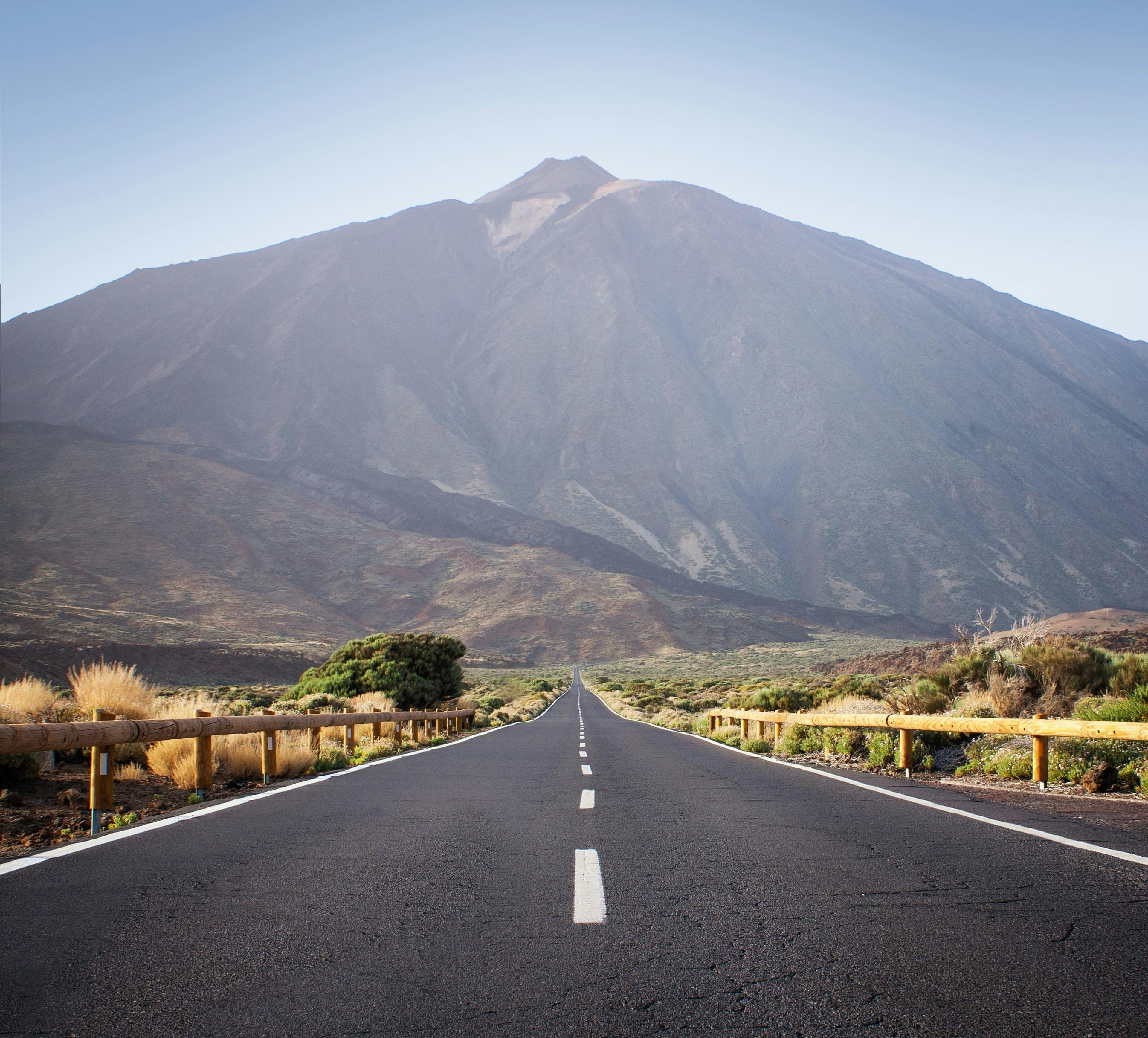 A road leads towards the imposing Mount Teide on Tenerife, framed by rugged terrain under a clear sky.