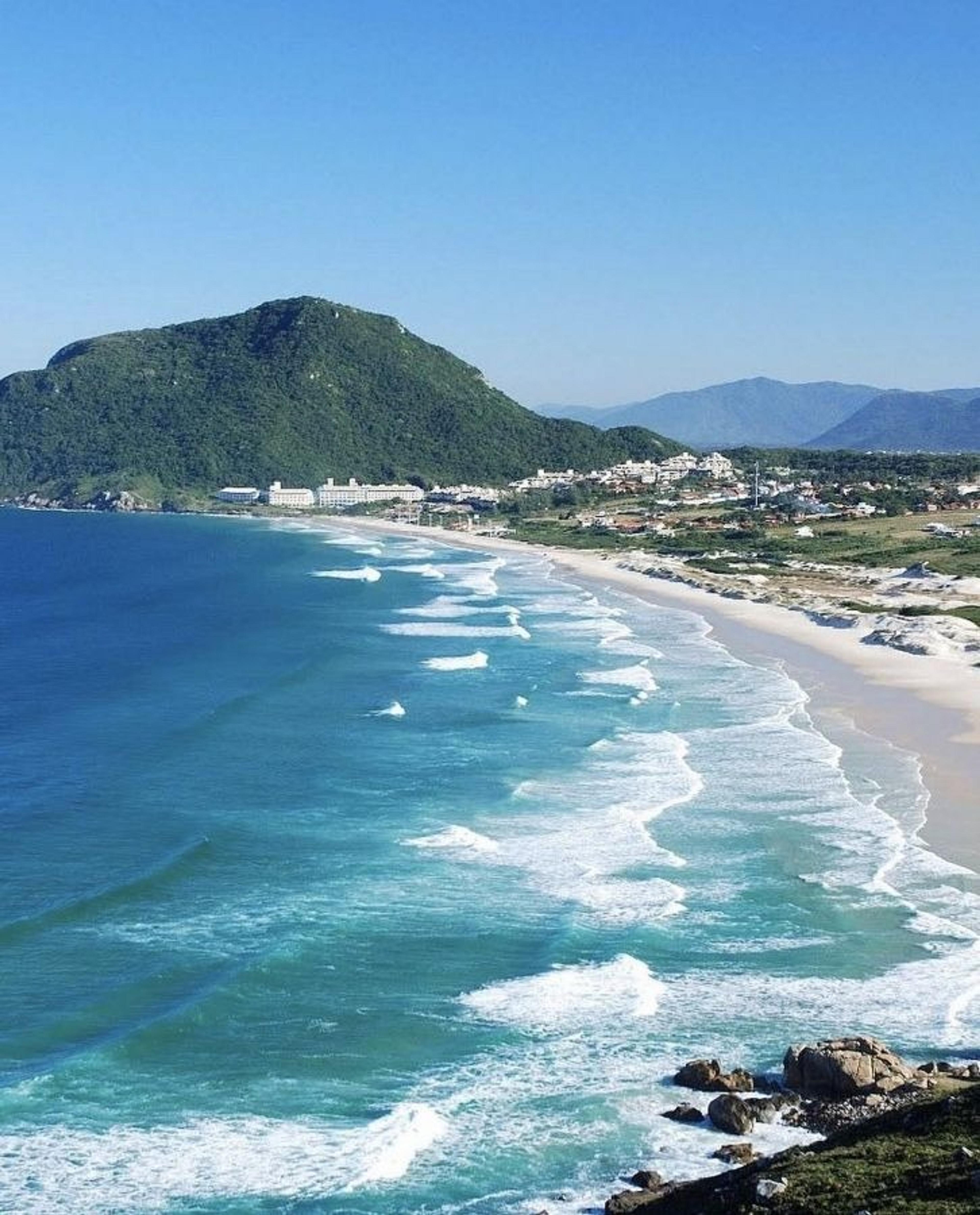 Vibrant waves crash onto the sandy shores of Praia do Santinho in Florianópolis, Brazil, with lush green hills looming in the background.