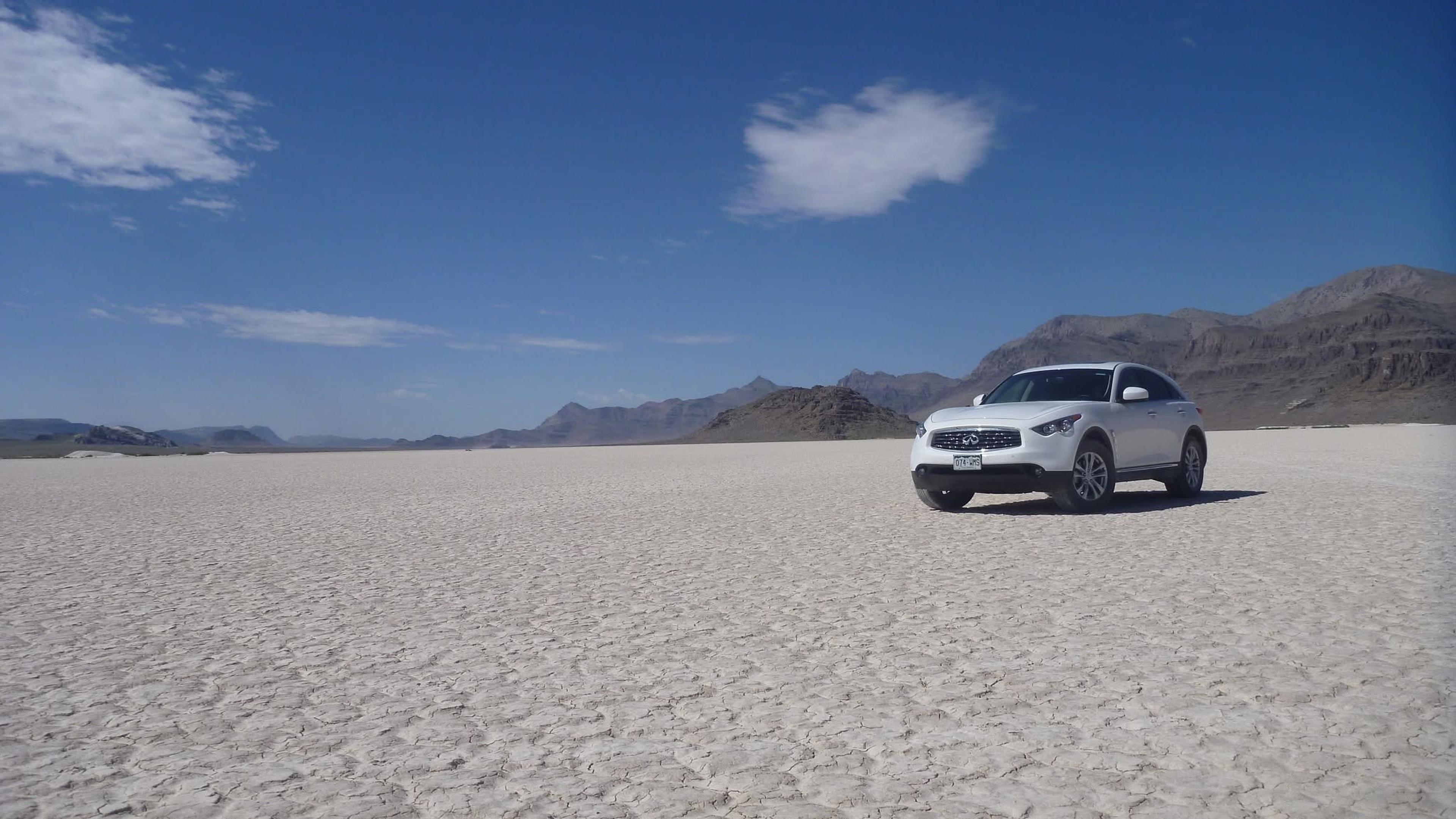 A white car stands on the vast, dried plain of the Bonneville Salt Flats in Utah, surrounded by distant mountainous terrain.