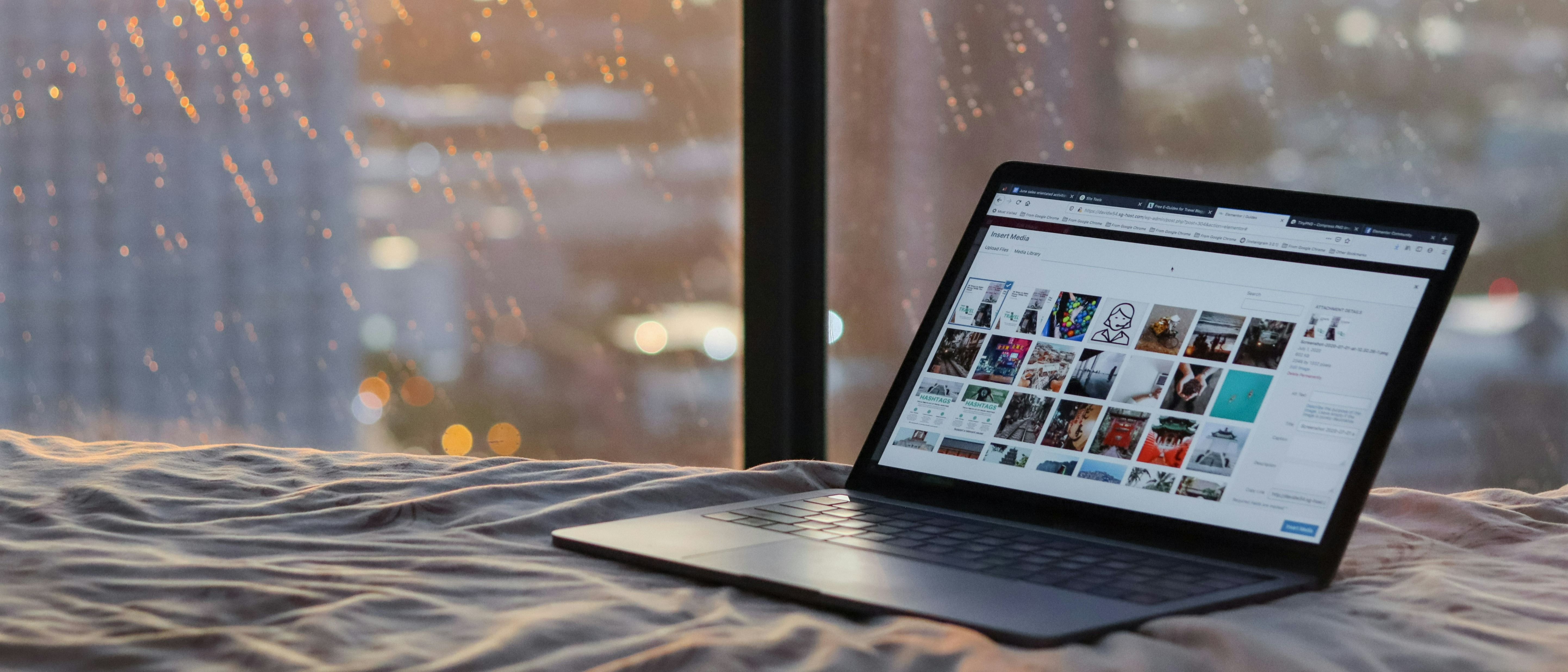 A laptop displaying a media gallery rests on a bed in front of a rain-speckled window with a cityscape visible in the background.