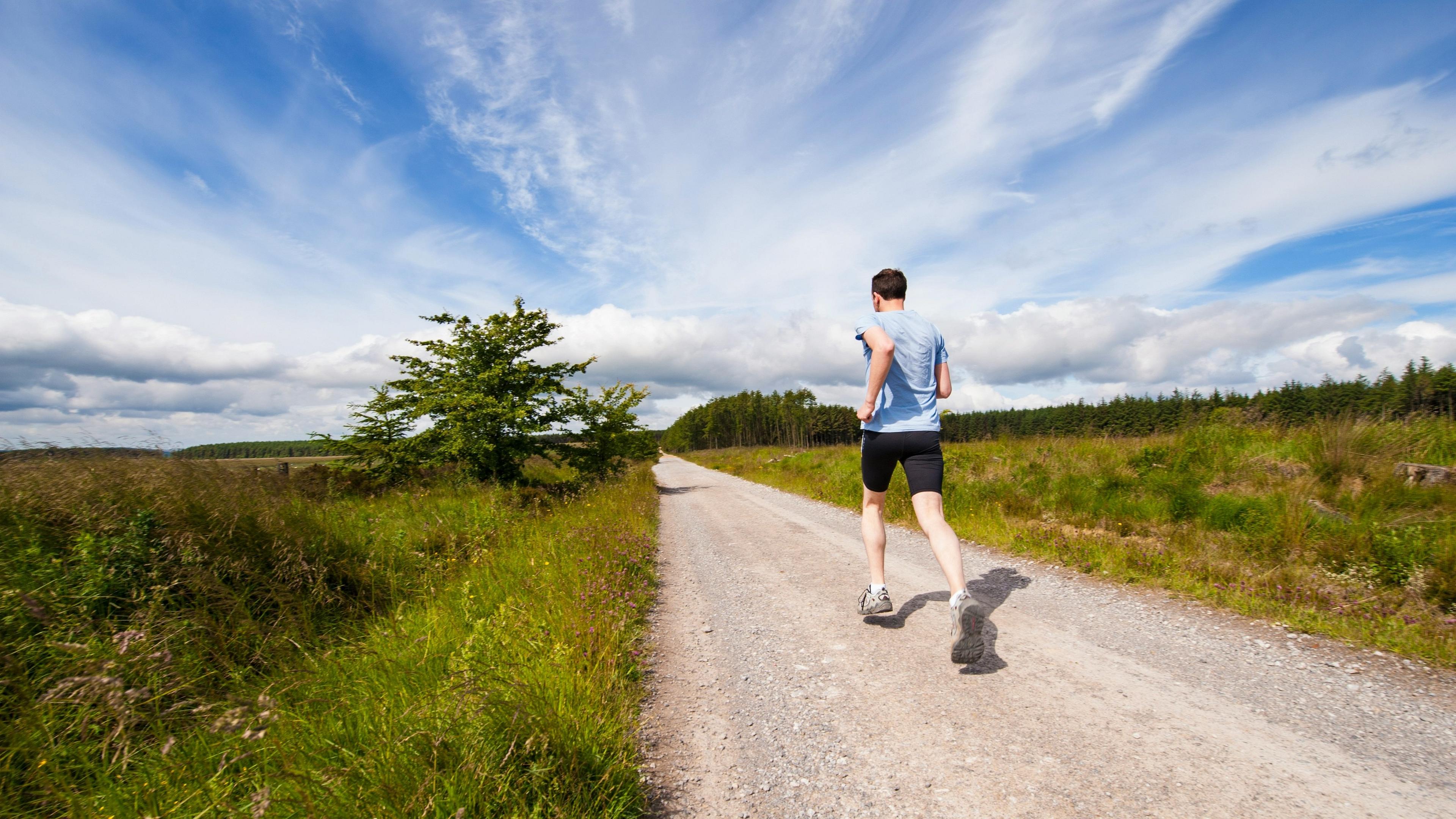 A person runs along a gravel path through lush greenery under a bright blue sky in a countryside setting.