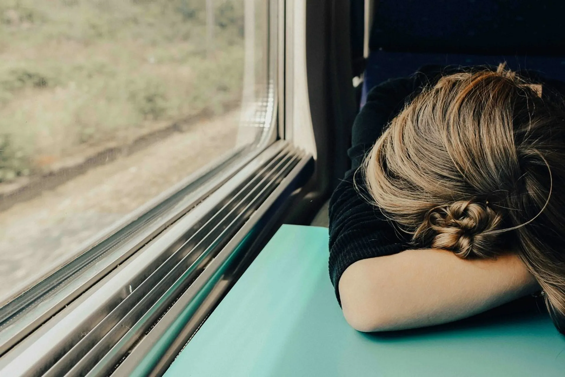 A person with their head resting on a table near a window on a moving train passes by a blurred countryside landscape.