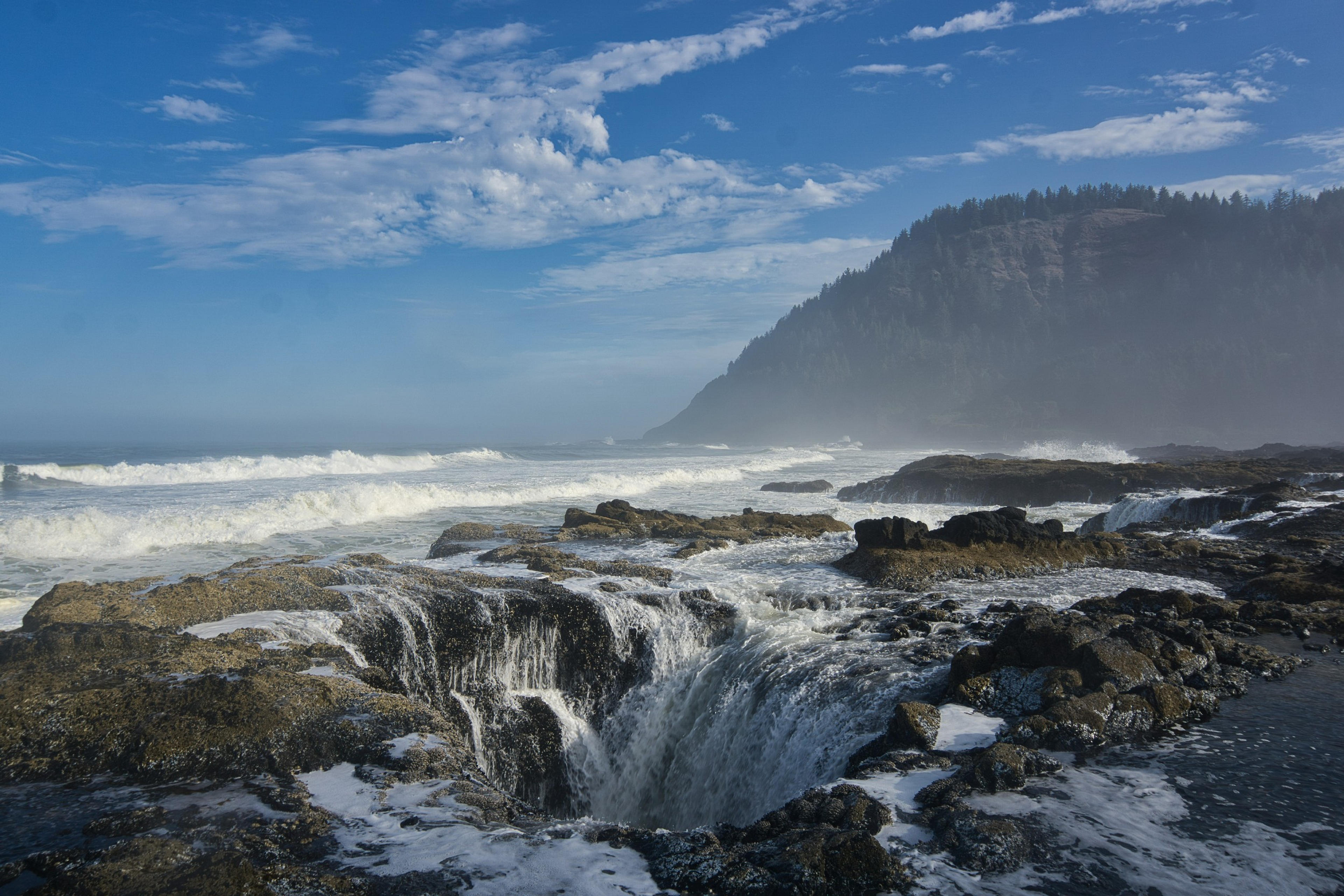 Waves cascade over the rocky Thor's Well along the Oregon coast, with a backdrop of misty hills and a vibrant blue sky.