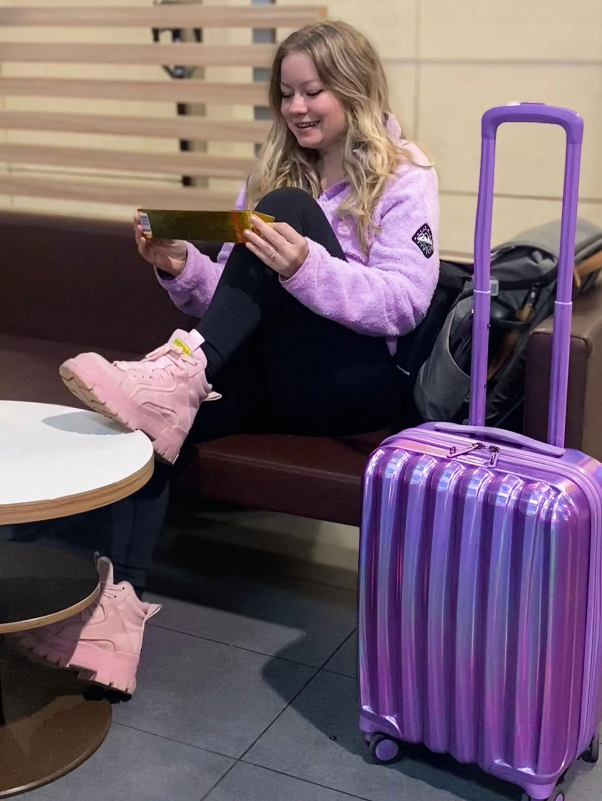 Flight Finder Kristi with her iridescent suitcase examines a Toblerone in an airport lounge dressed in purple.