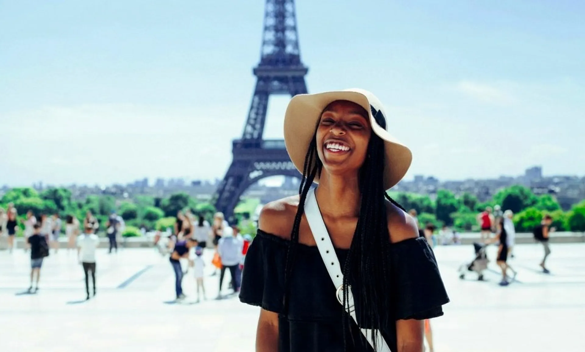 A woman wearing a hat stands smiling in front of the Eiffel Tower
