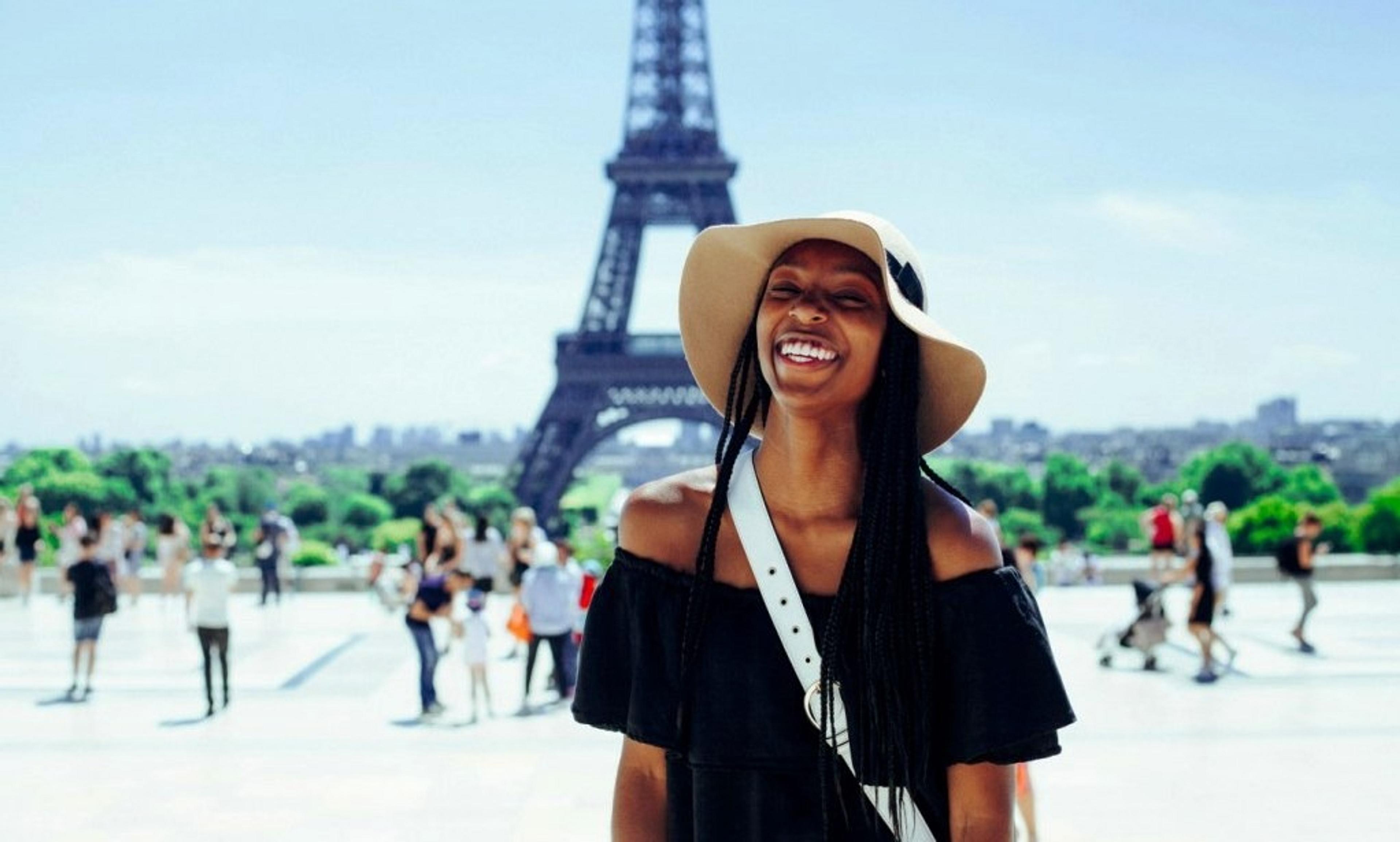A woman wearing a hat stands smiling in front of the Eiffel Tower