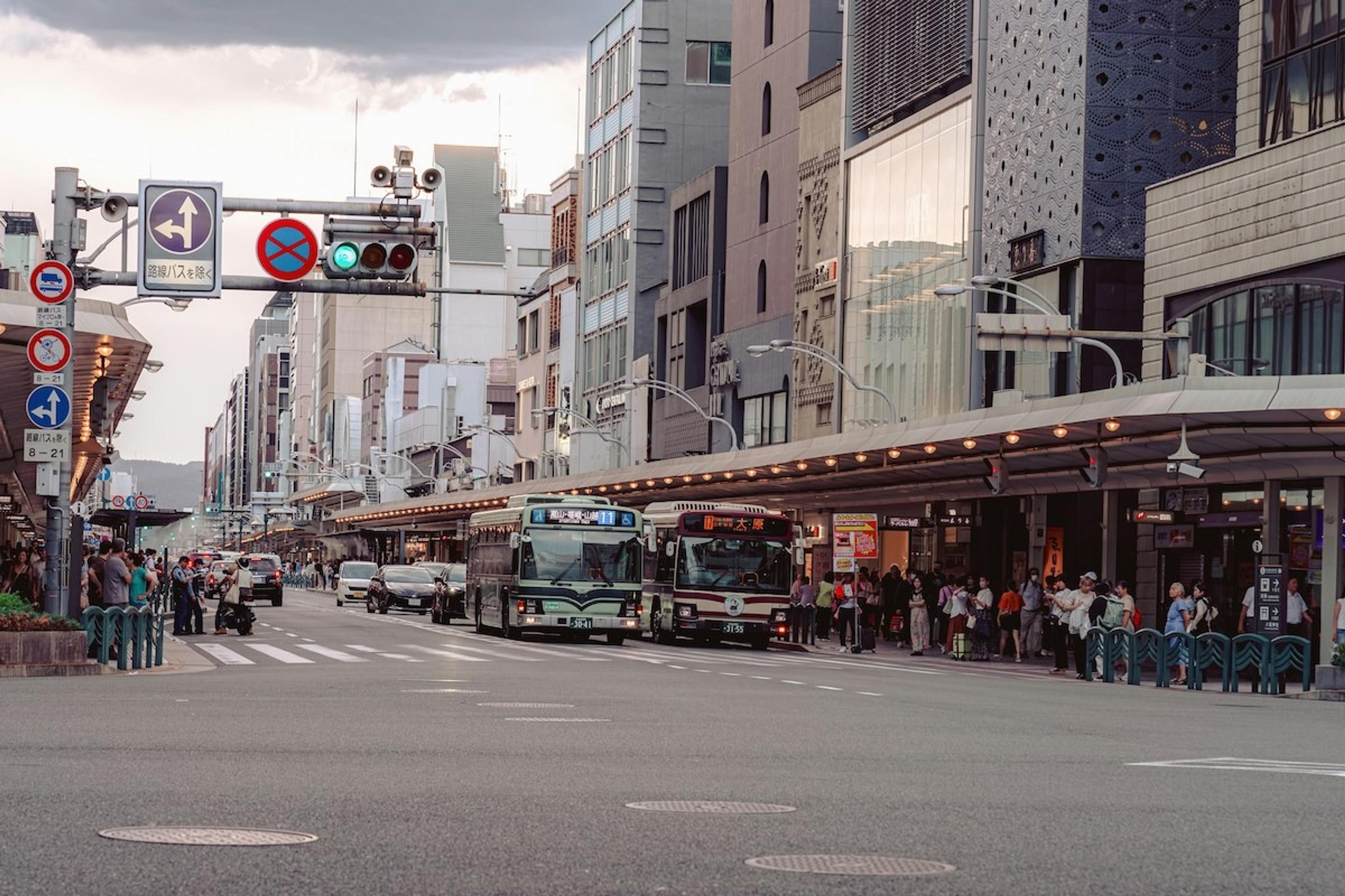 Kyoto's bustling Shijo Street features city buses, modern buildings, and a busy pedestrian sidewalk.