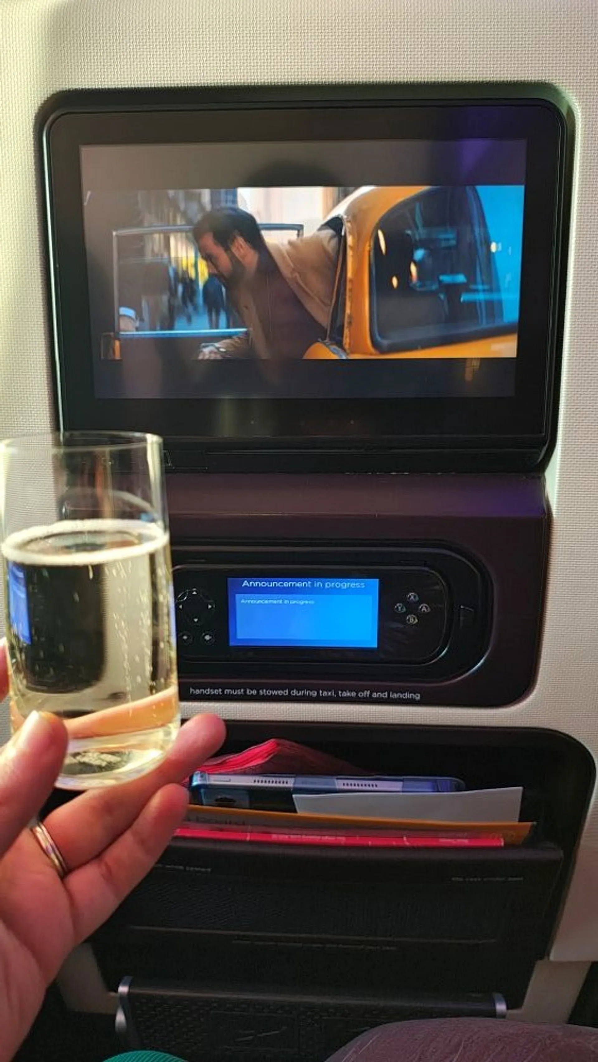 An airplane passenger enjoys a drink while watching a movie featuring a yellow taxi on their in-flight entertainment screen.